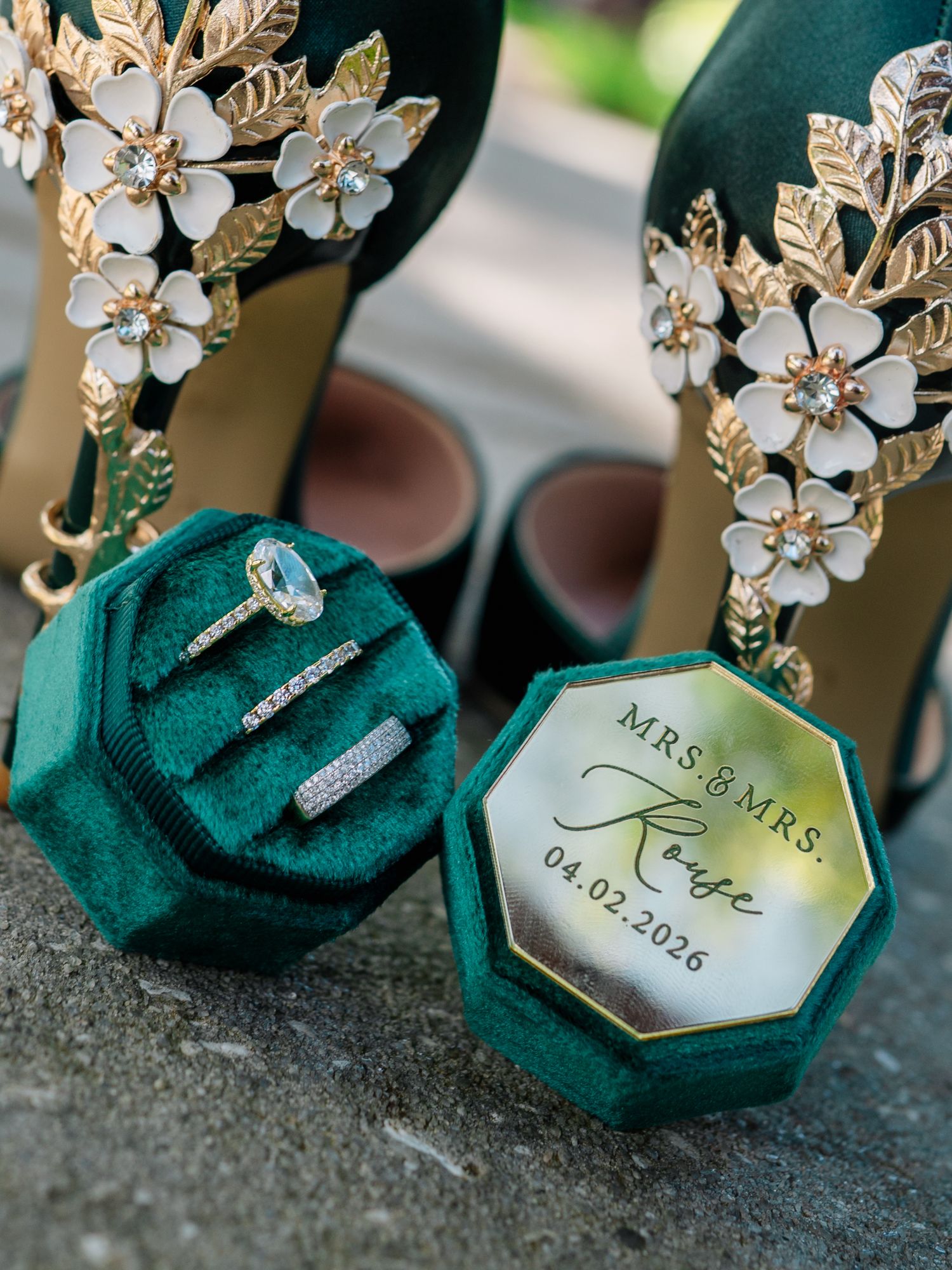 Green velvet ring box with Mrs. & Mrs. Rouse wedding bands beside emerald heels adorned with gold floral embellishments.