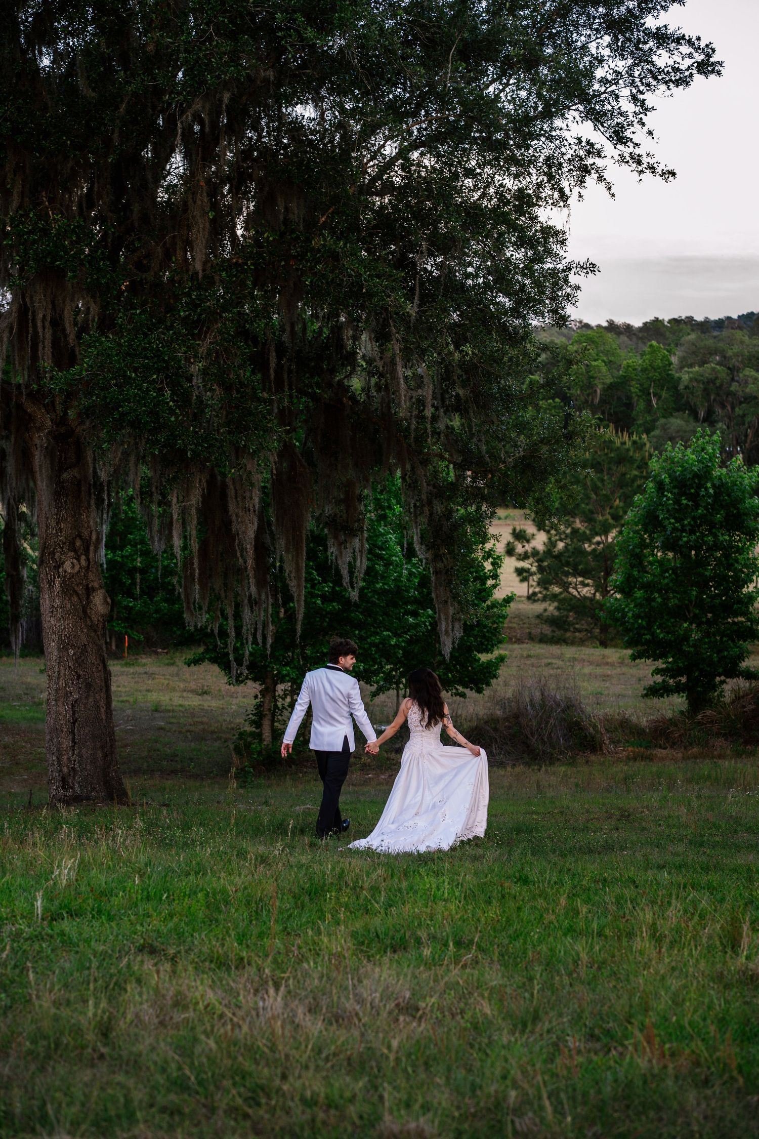 A bride and groom walk hand in hand through a lush green field surrounded by moss-draped oak trees at dusk.