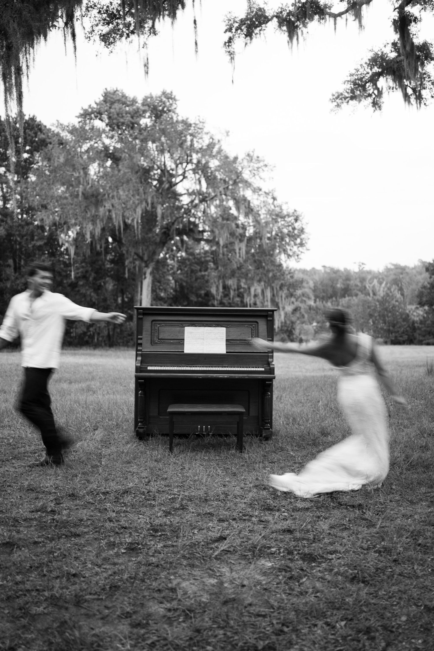 Black and white photo of a couple dancing around an upright piano outdoors under Spanish moss-draped trees.