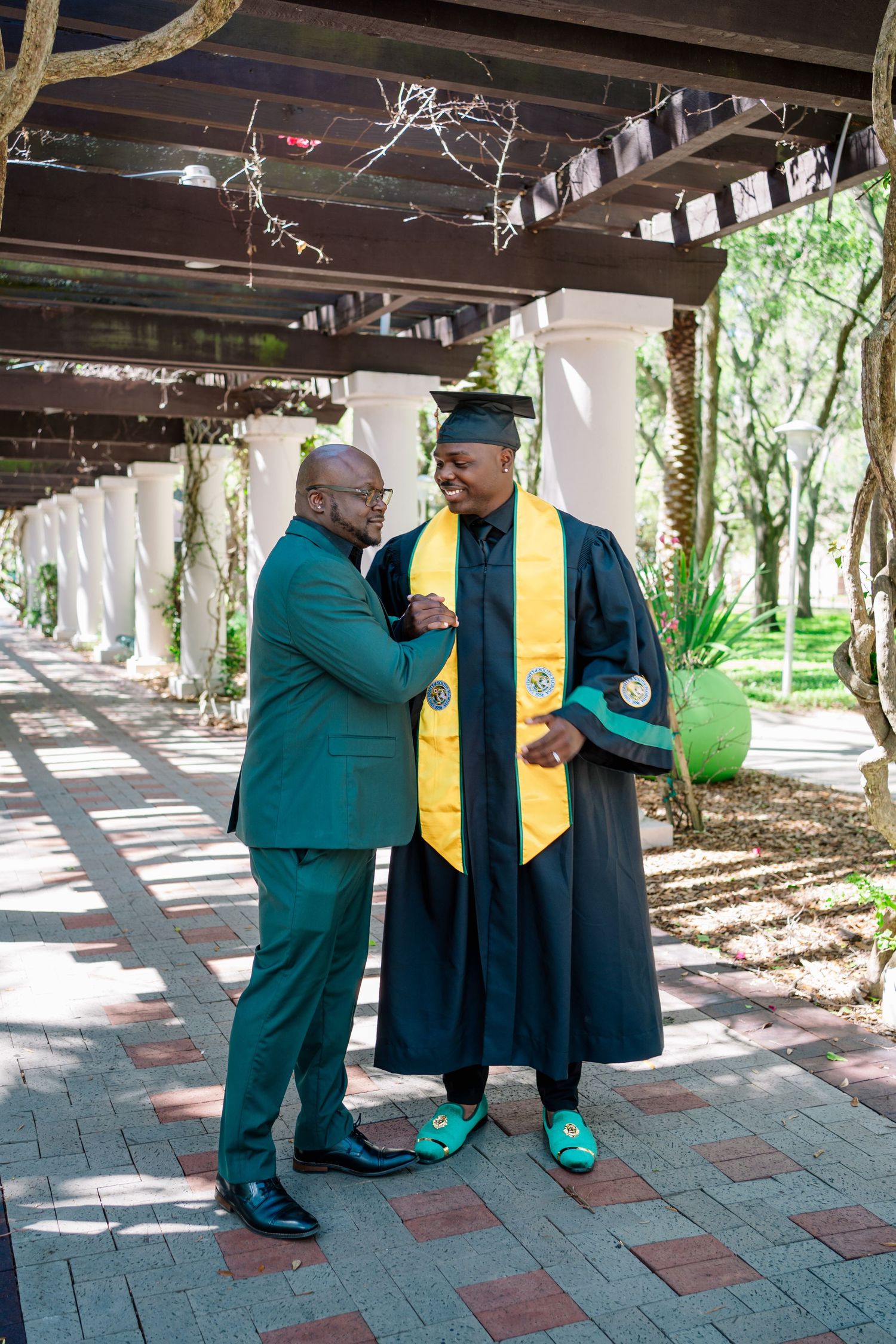 Graduate in cap and gown with gold stole celebrates with proud family member in green suit at outdoor ceremony.