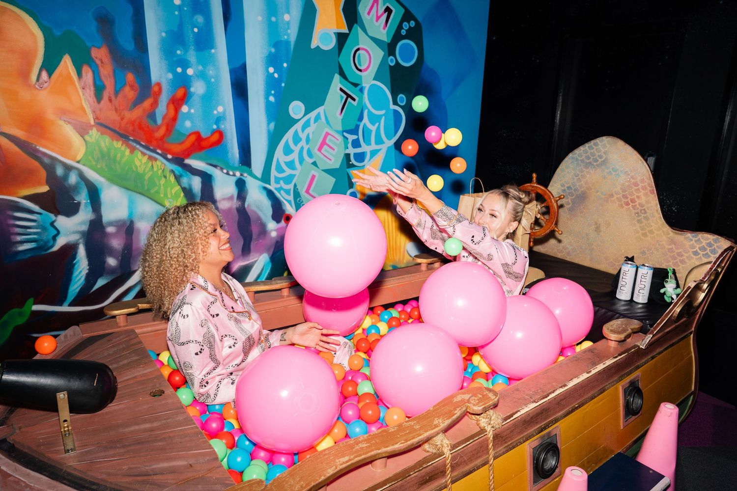 Two young girls playing with pink balloons in a decorative boat at a colorful mermaid-themed party.