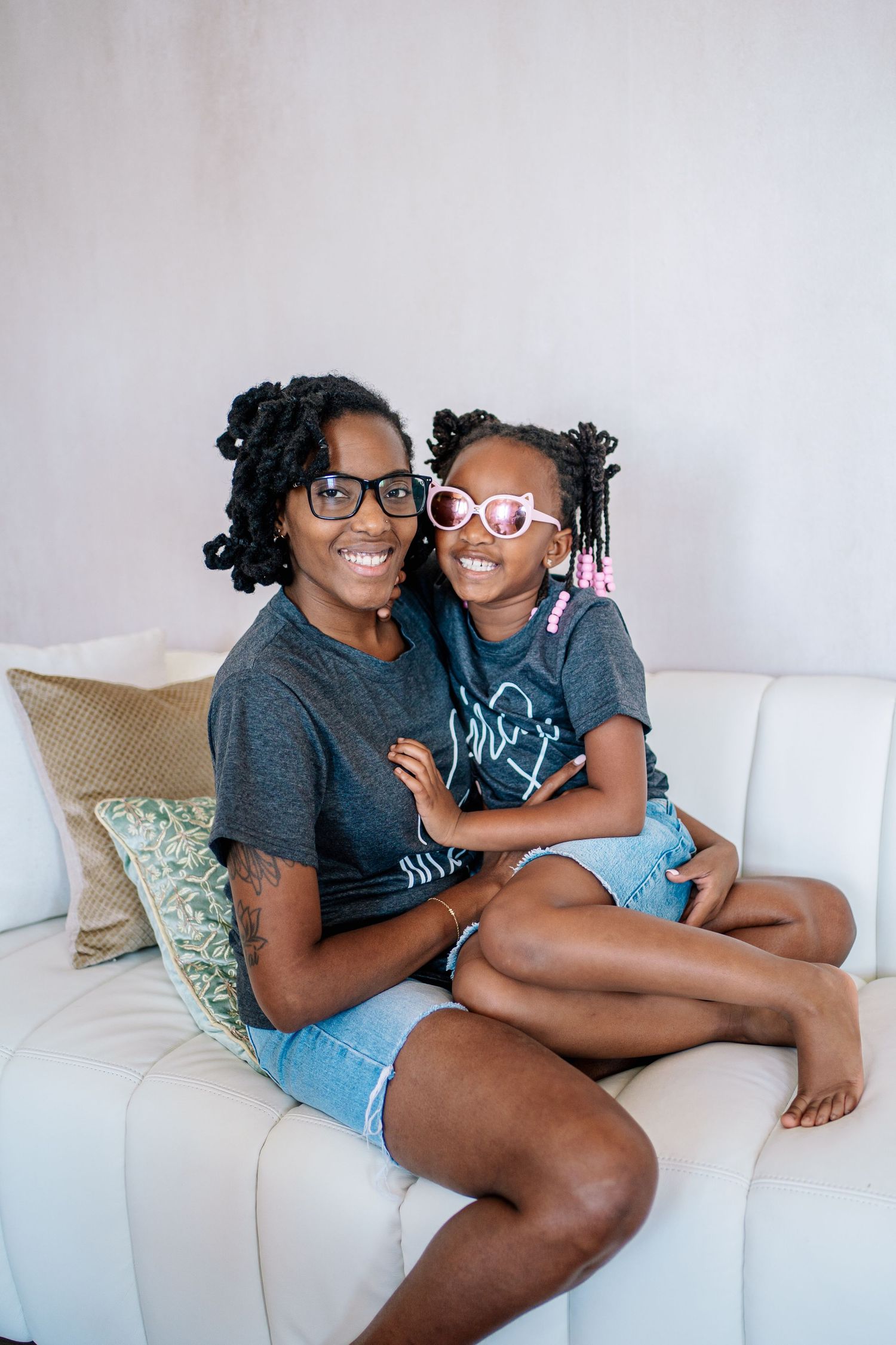 A smiling woman and young girl in matching gray shirts and denim shorts sit together on a white couch.