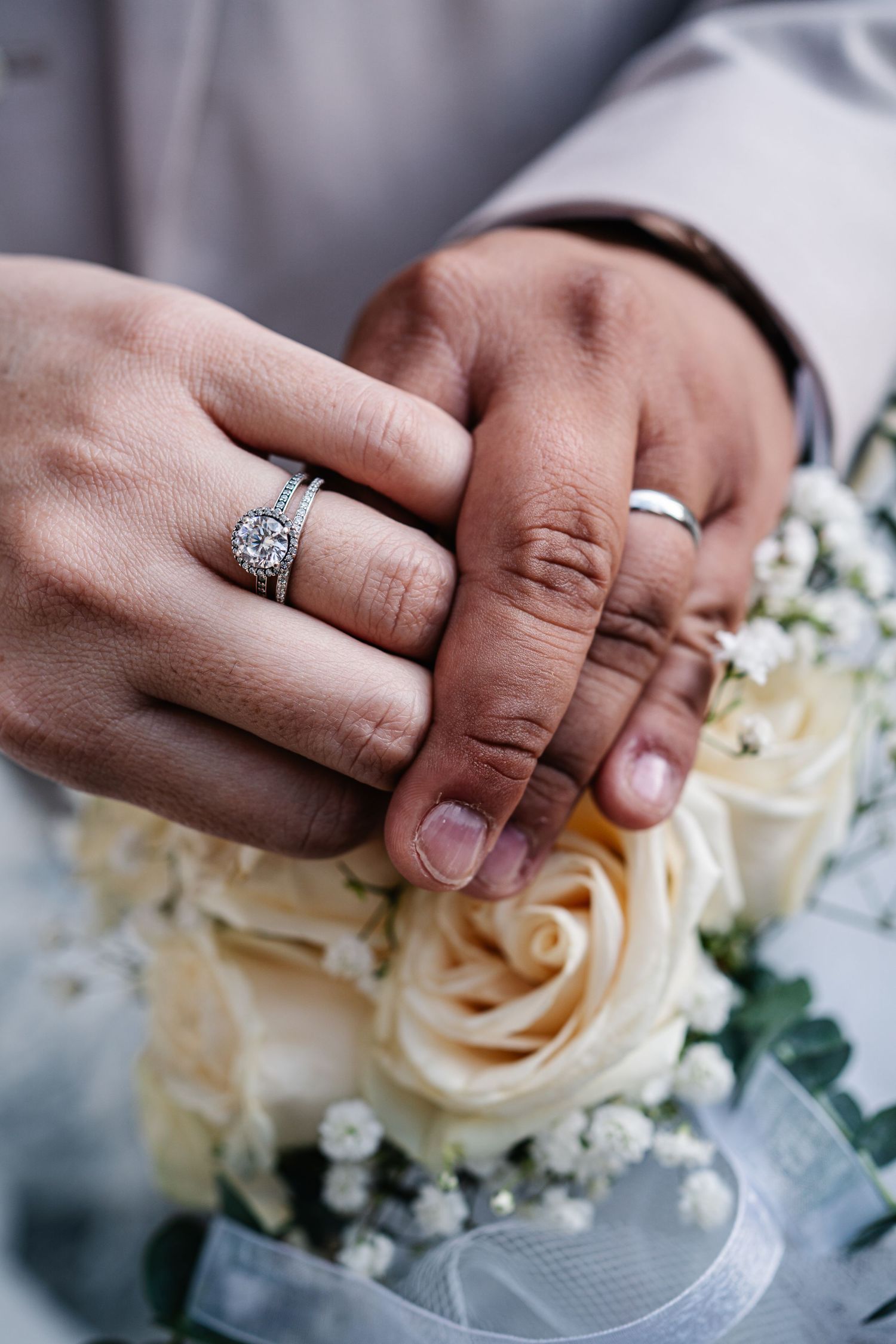 Close-up photo of wedding rings on hands with cream roses in the background showing romantic details.