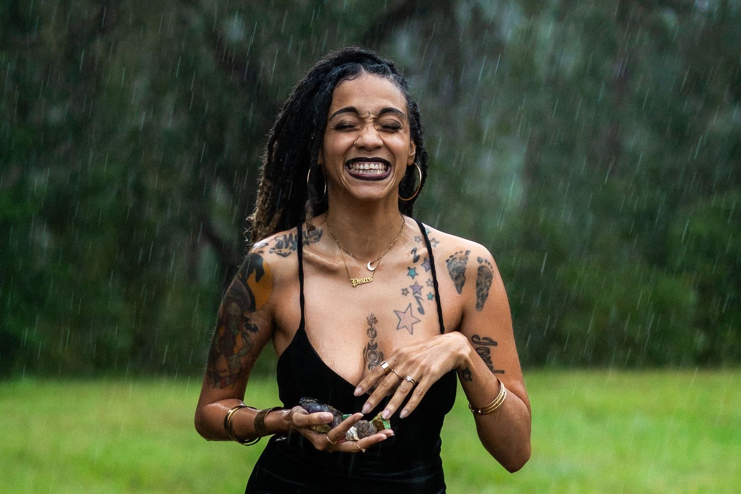 A person in a black tank top laughs joyfully while standing in the rain on a green lawn.