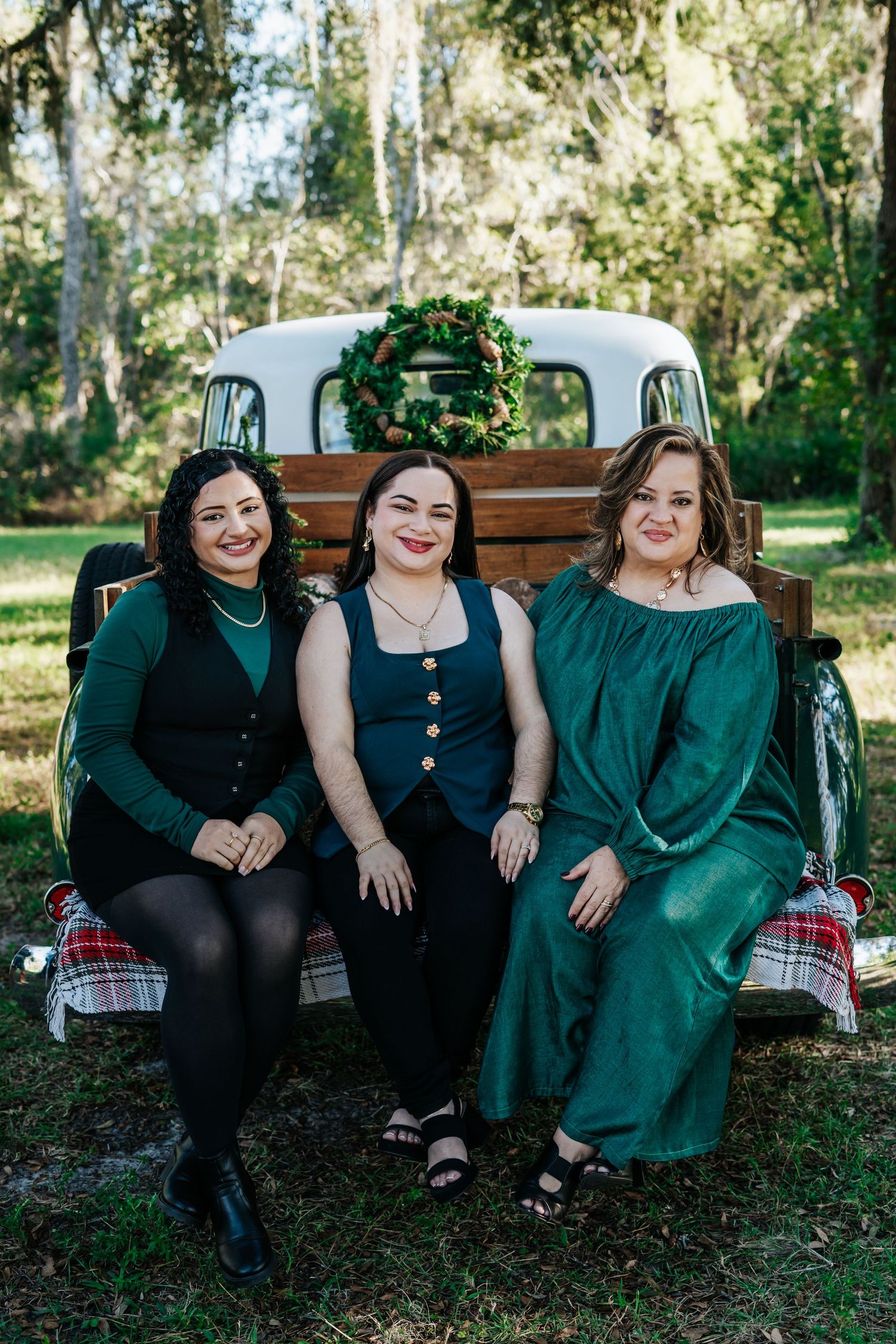 Three friends in coordinating green outfits sit together in the back of a vintage truck with a holiday wreath.