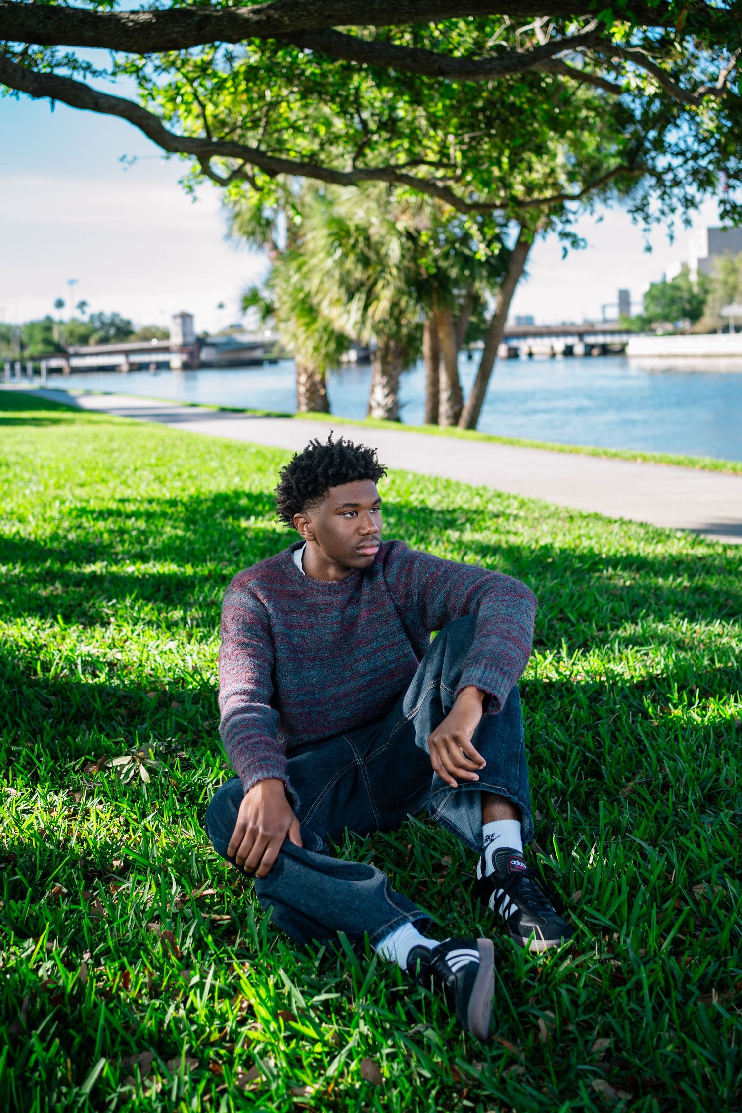 Young Black man in gray sweater and jeans sits on lush green grass by waterfront under shady trees.