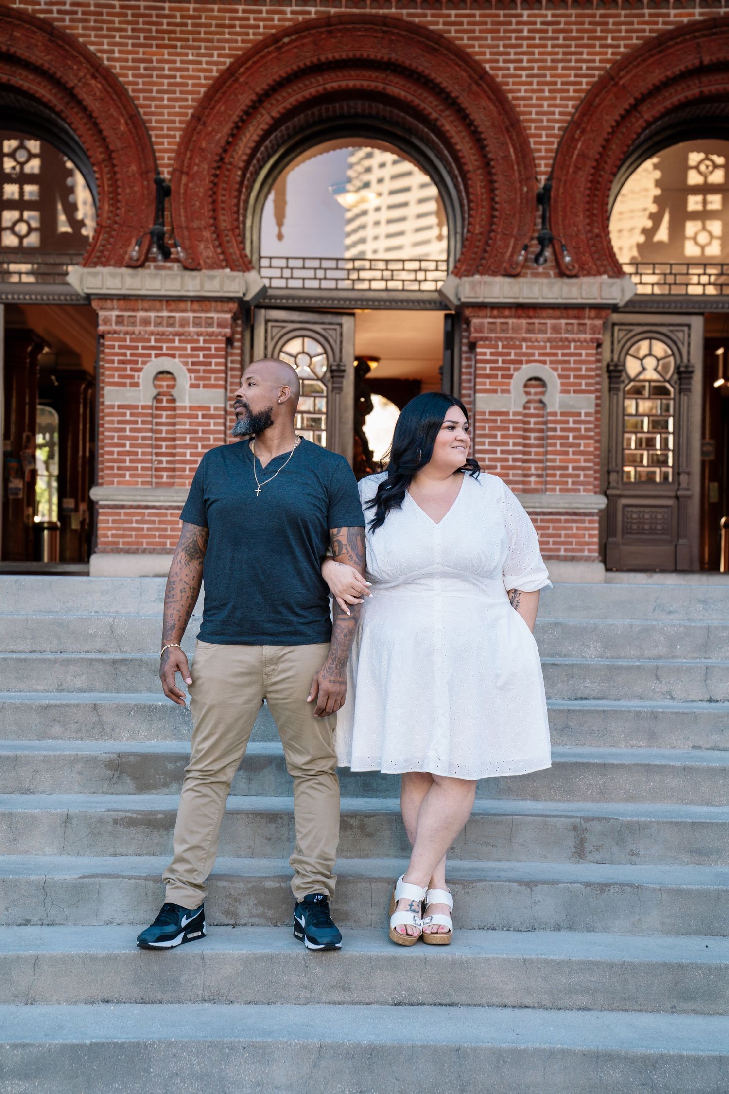 Couple posing on steps of a red brick building, man in khakis and woman in white dress, engagement photo.