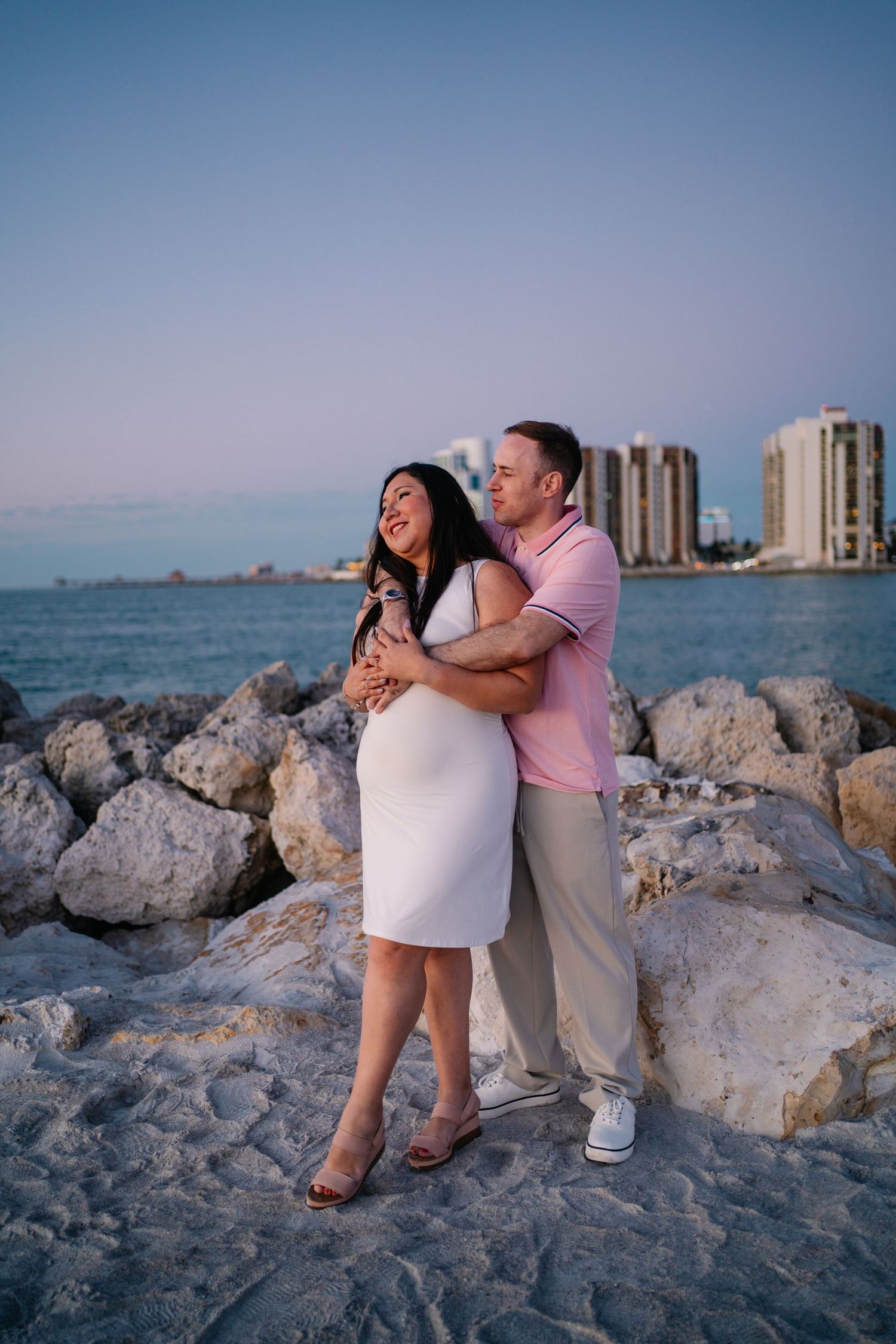 A couple embraces on rocky shoreline at sunset, woman in white dress, man in pink polo, with ocean and city skyline behind.