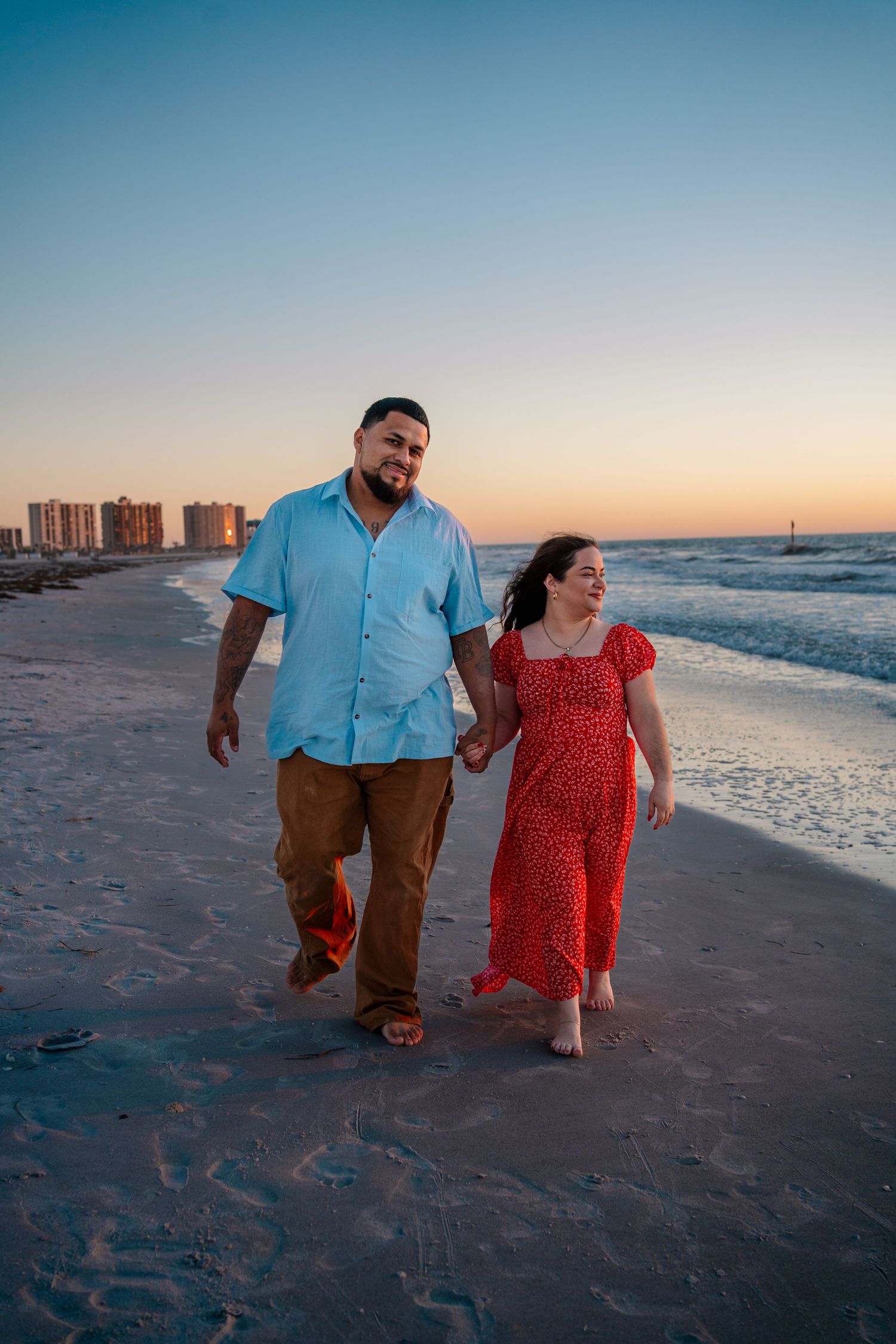Couple holding hands walking on beach at sunset, man in blue shirt and woman in red floral dress.