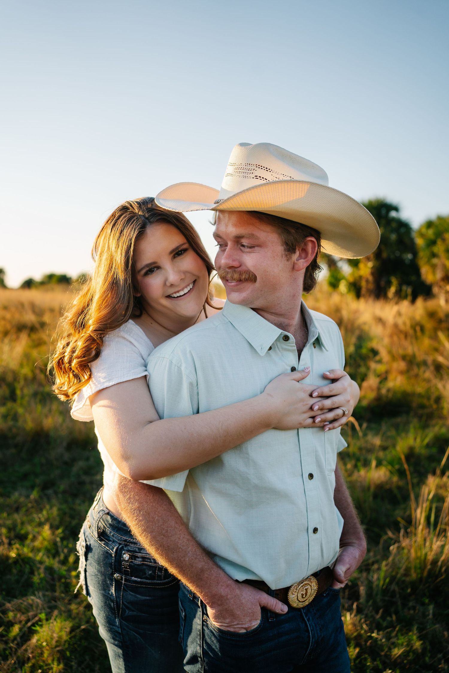 A smiling couple in cowboy hats embrace in a golden sunlit field, wearing jeans and casual shirts.