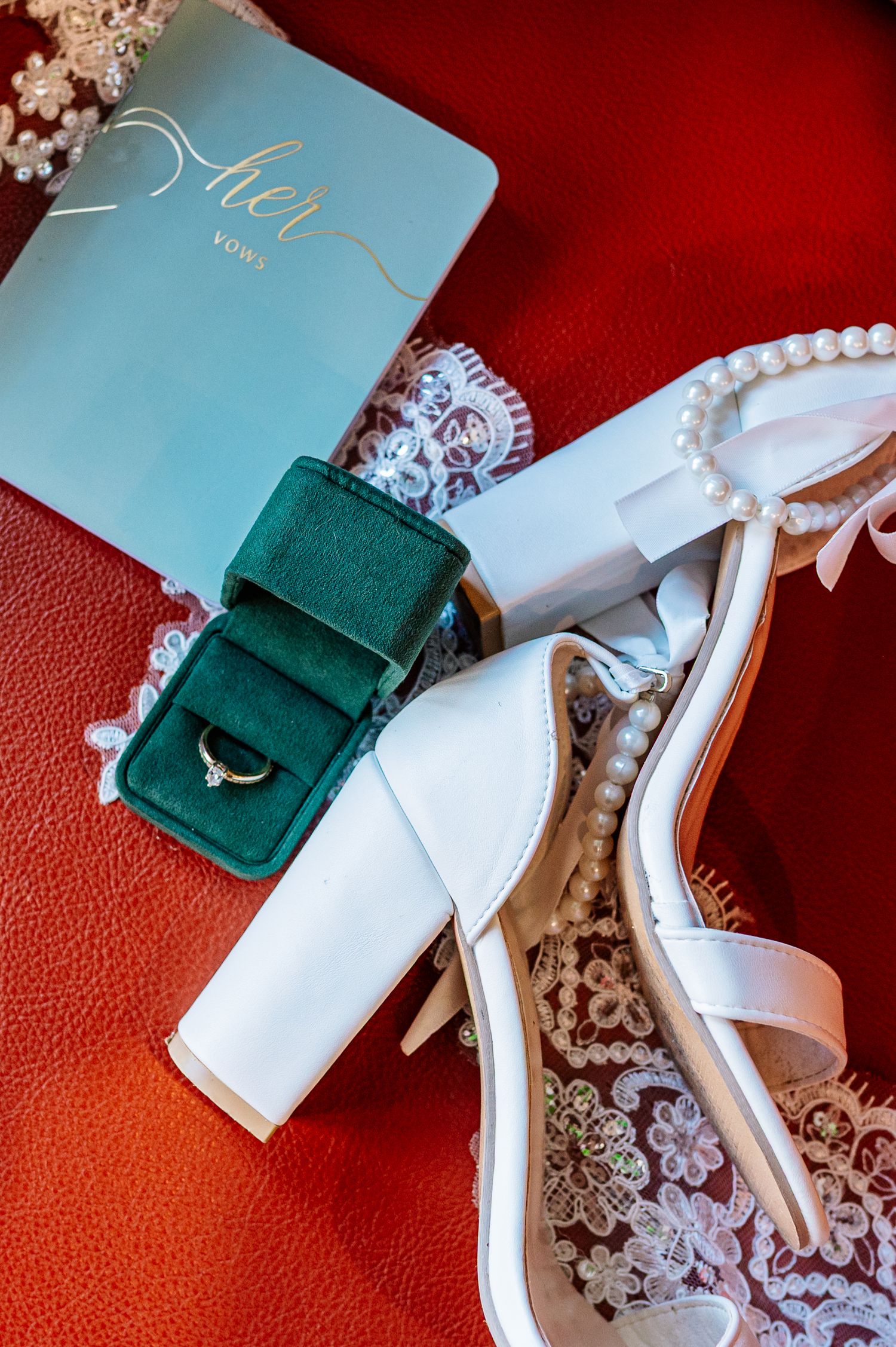 White bridal heels with pearl accents beside a green velvet ring box and light blue vow book on red background.