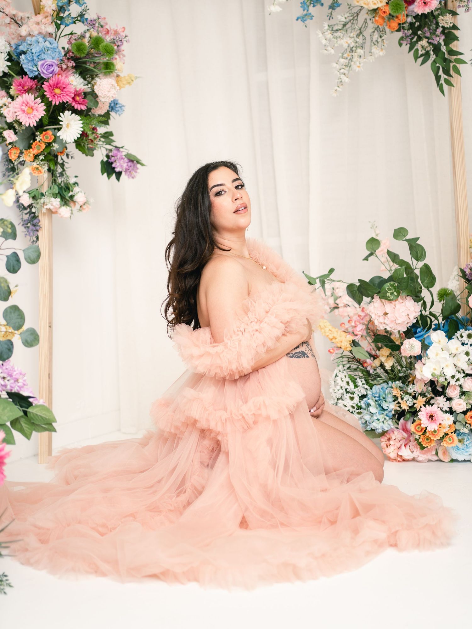 Pregnant woman in pink ruffle tulle gown poses gracefully among colorful floral arrangements in a bright studio setting.