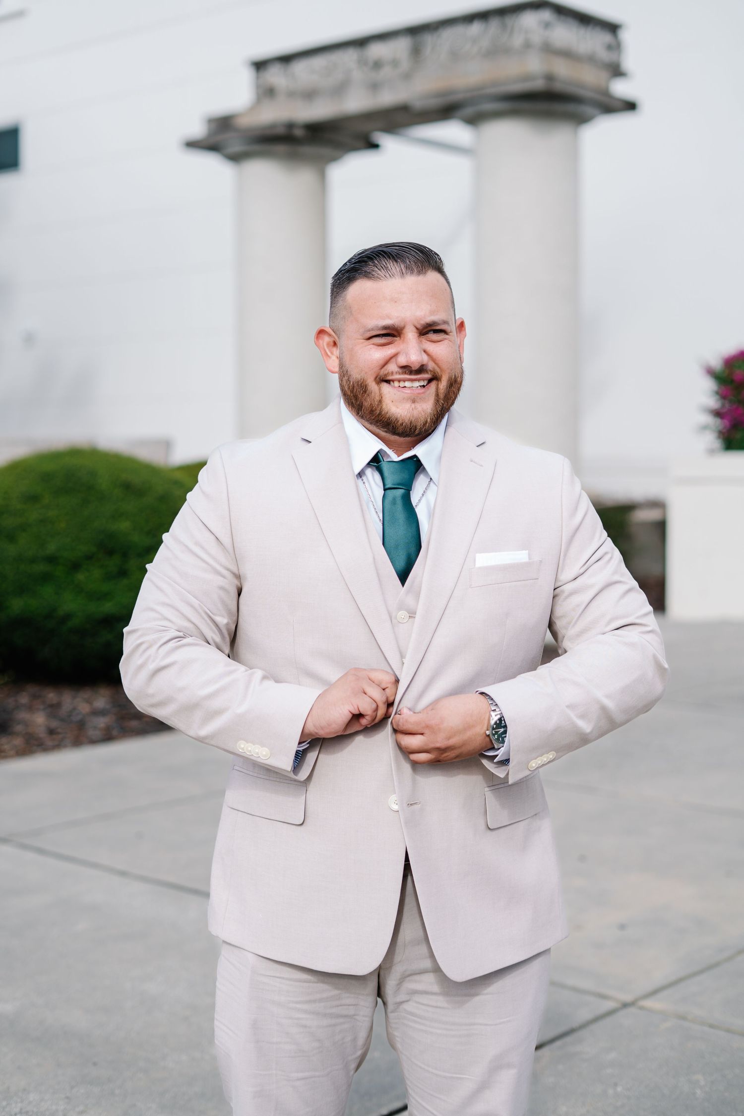 A person in a light beige suit and teal tie poses outdoors near a white building.