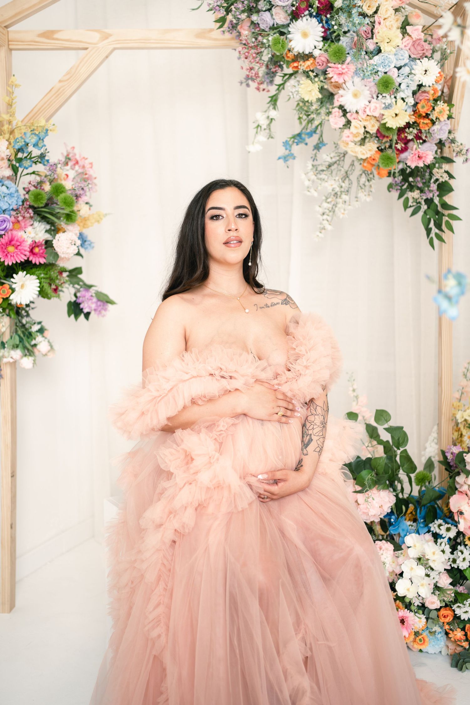 Pregnant woman in blush pink ruffle tulle gown poses gracefully among colorful floral arrangements in a bright studio.