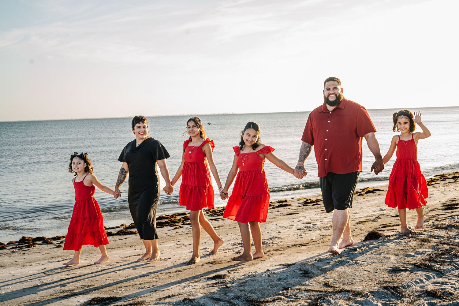Family of six walking hand-in-hand on a beach at sunset, wearing coordinated red and black outfits.