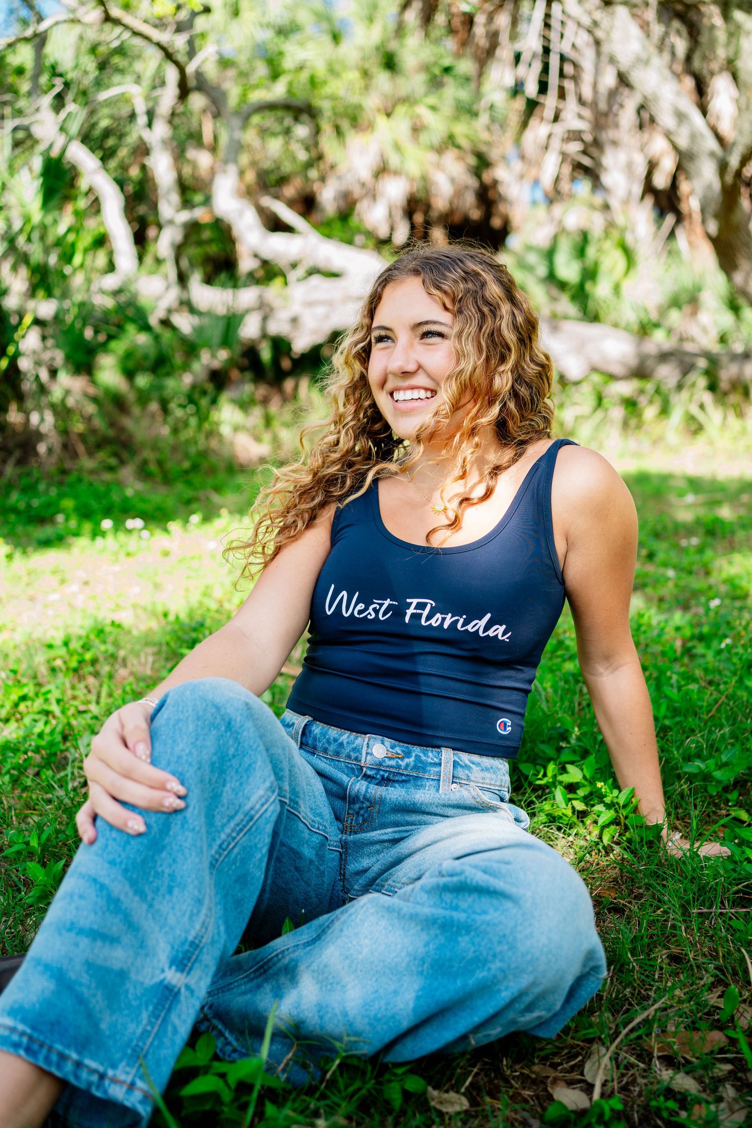 Smiling young woman with curly hair sitting on grass wearing a navy West Florida tank top and wide-leg jeans outdoors.