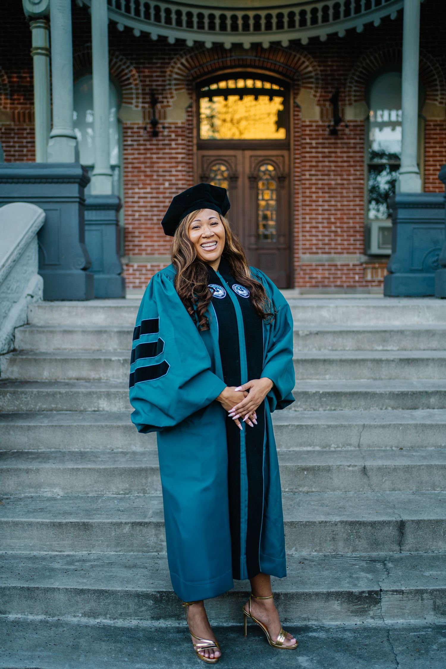 Smiling graduate in teal doctoral gown and black cap poses on stone steps outside brick building.