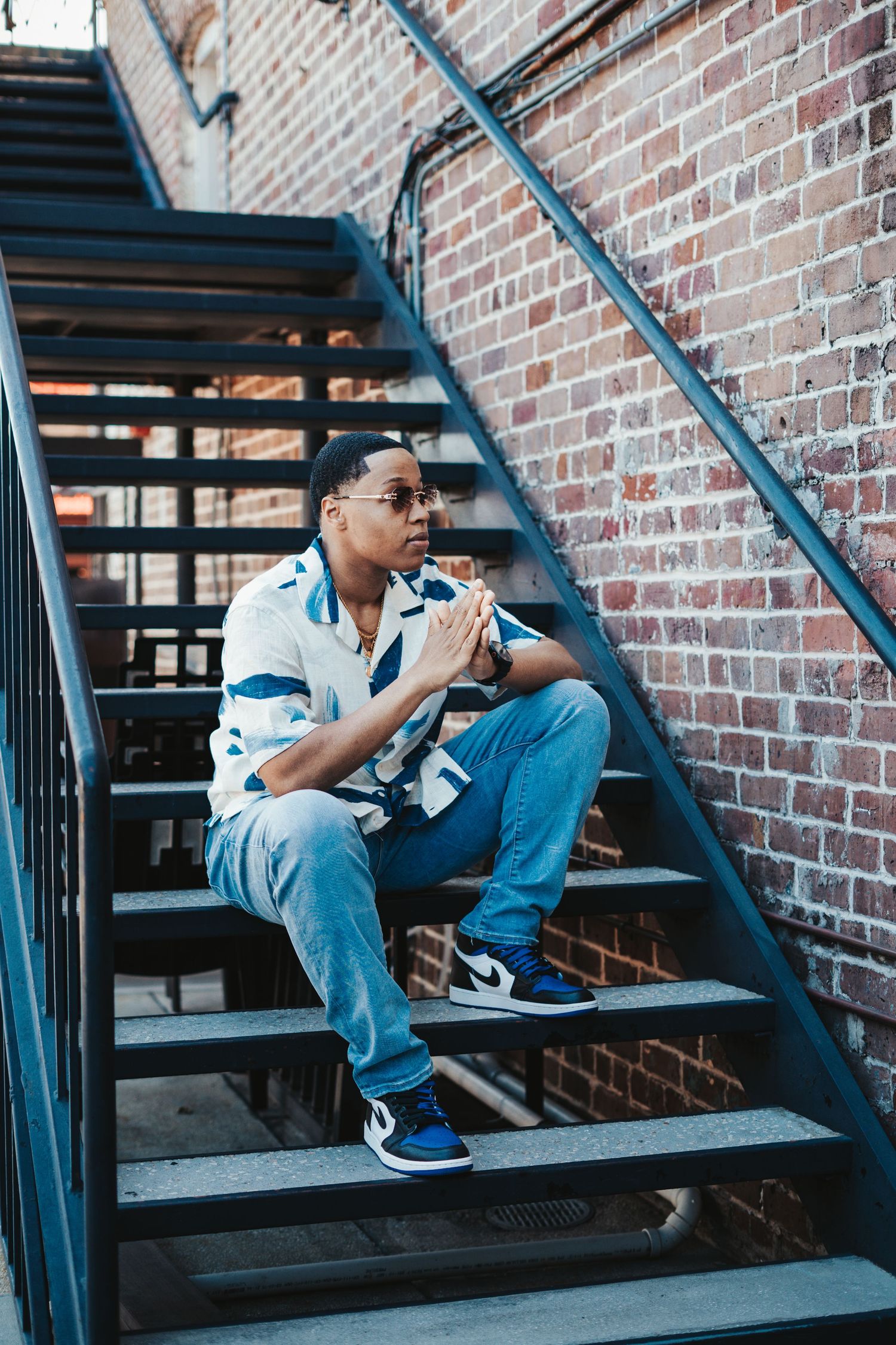 Person in blue denim sits contemplatively on metal fire escape stairs against brick wall background.