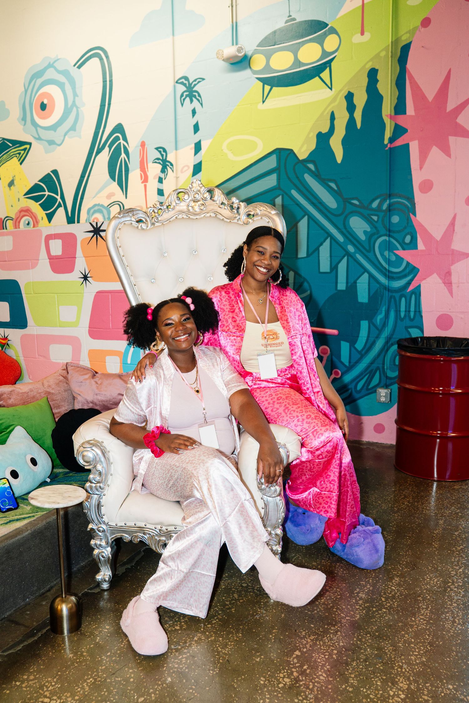 Two women in colorful pajamas pose on a silver throne chair in front of a vibrant retro-style mural.
