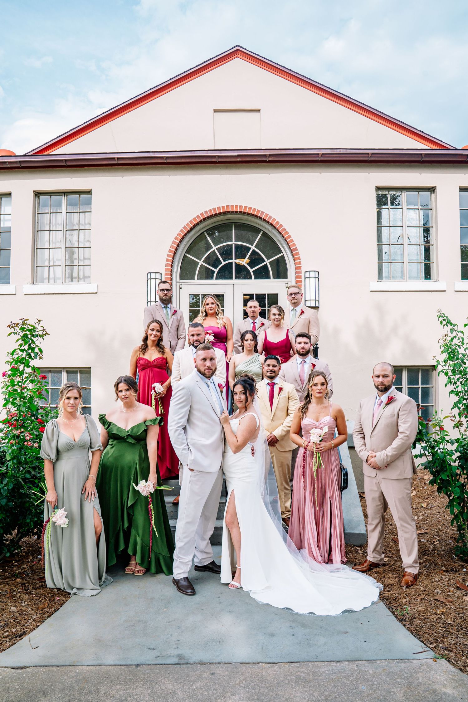 Wedding party poses in front of elegant venue, bride and groom centered with bridesmaids in green, pink, and red gowns.