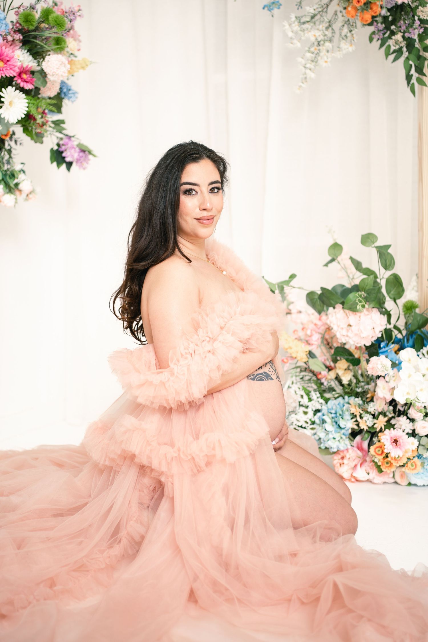 Pregnant woman in pink tulle robe poses gracefully among colorful flowers in a bright studio maternity photo session.