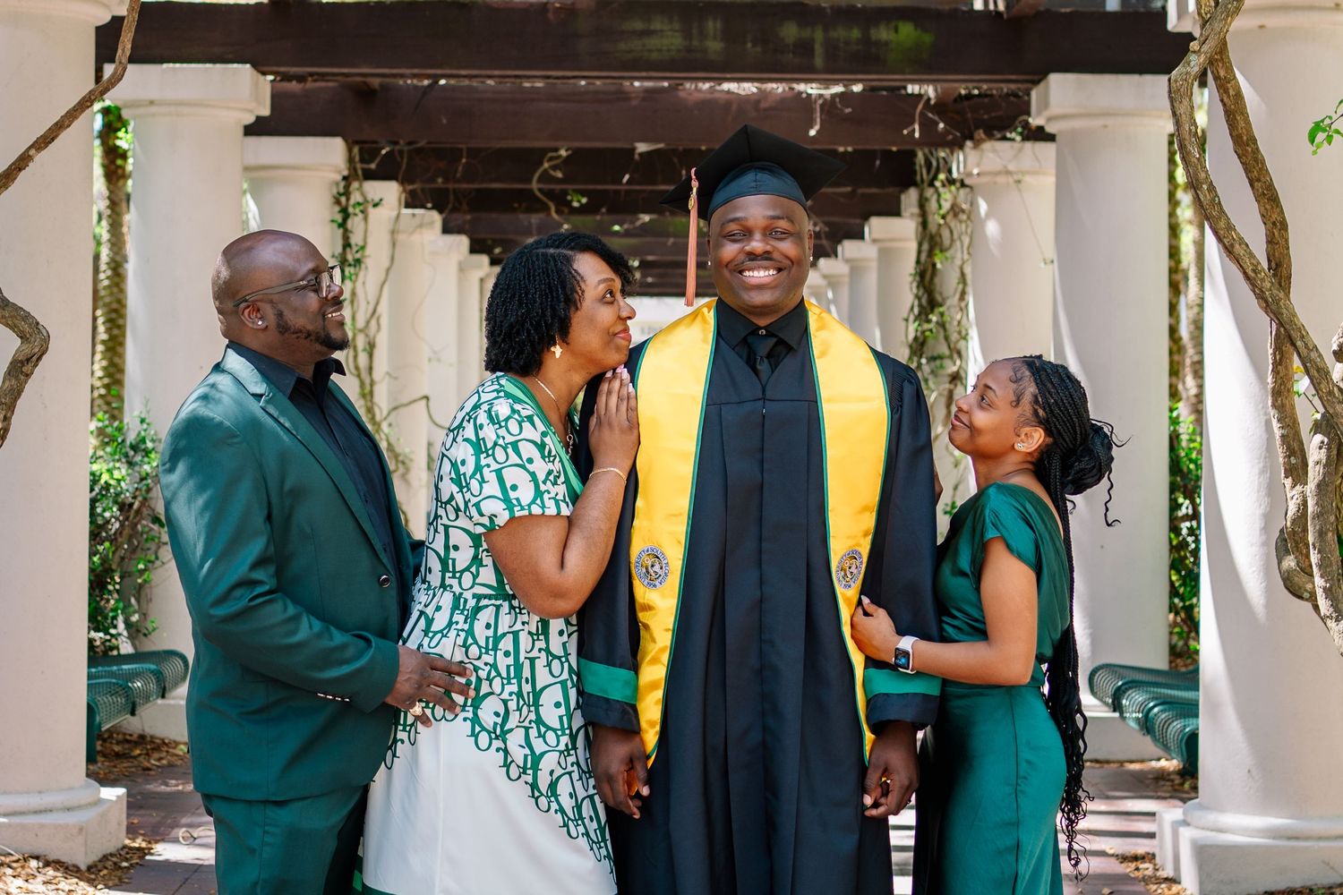 Graduate in cap and gown smiles with proud family members in green outfits at outdoor graduation celebration.
