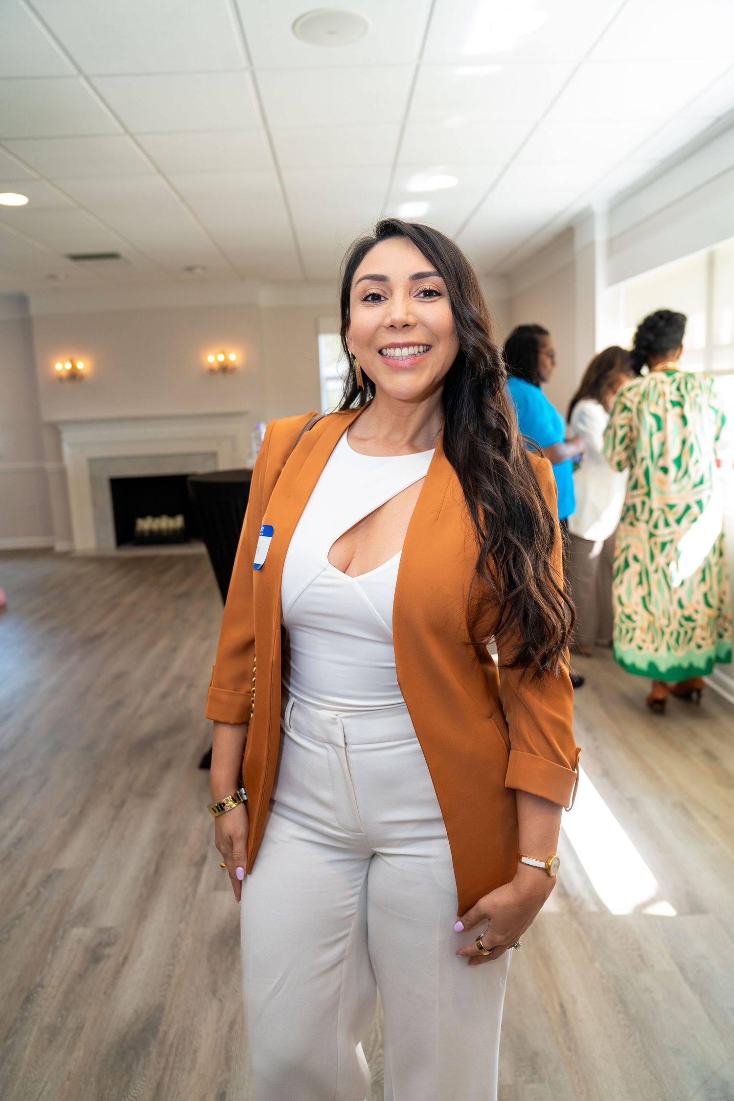 Woman in orange blazer and white outfit smiling at indoor networking event.