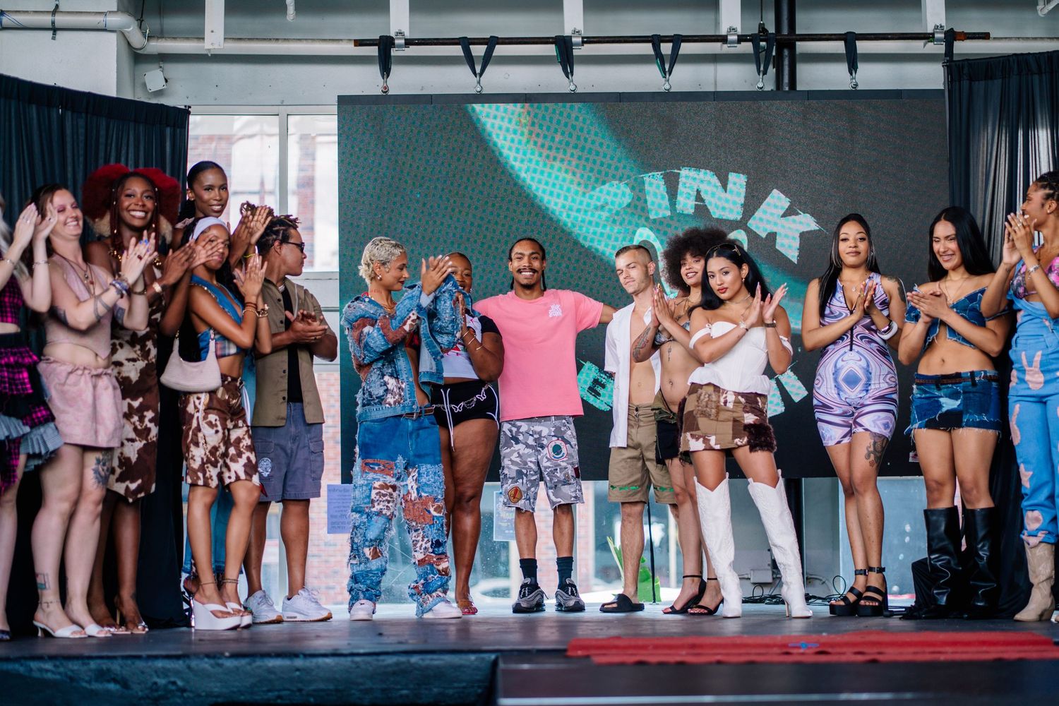 Diverse group of models and designers celebrate on a runway stage at a colorful fashion show event.