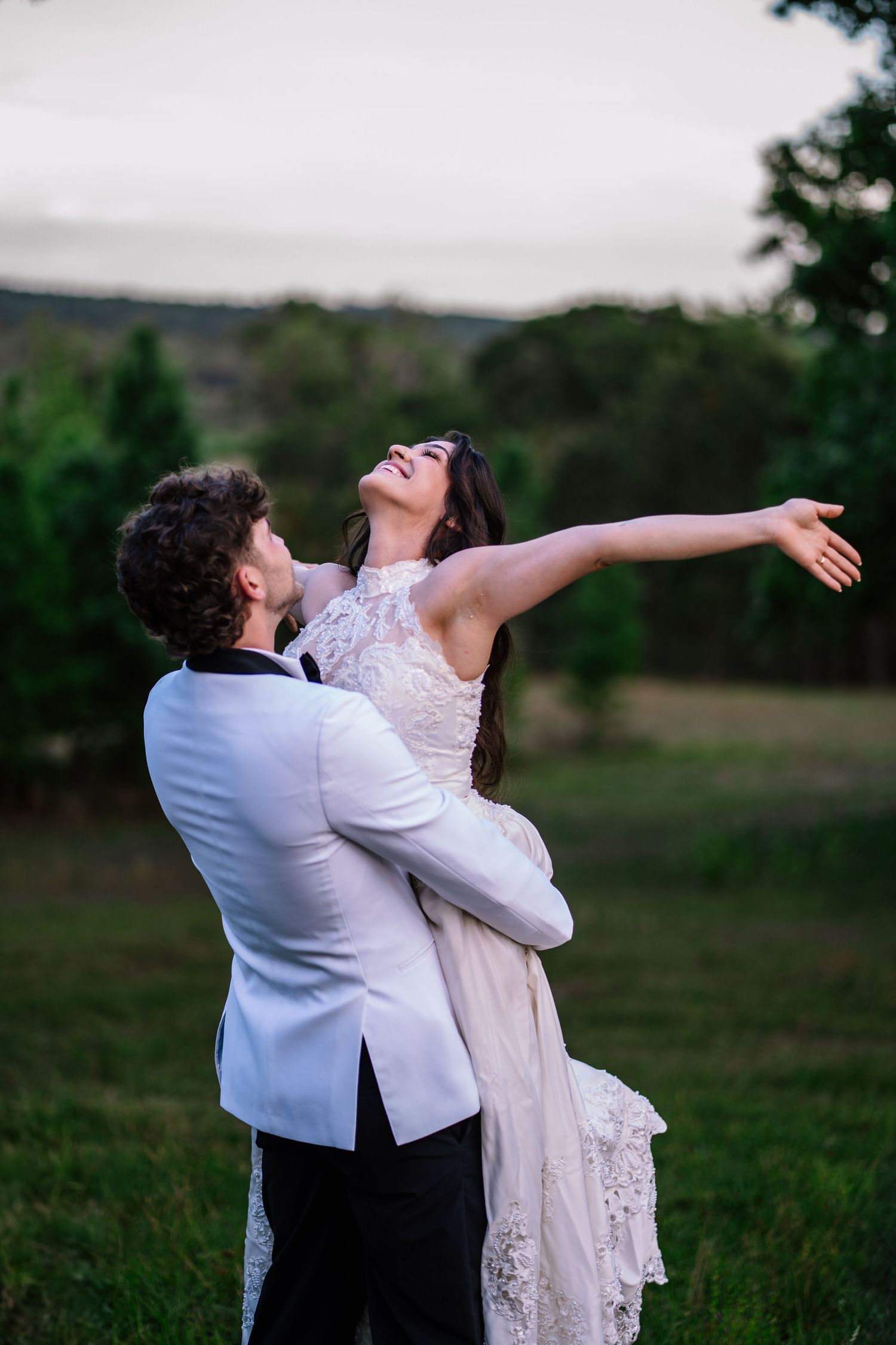 Groom in white tuxedo lifting joyful bride in lace wedding dress outdoors on green grass at dusk.