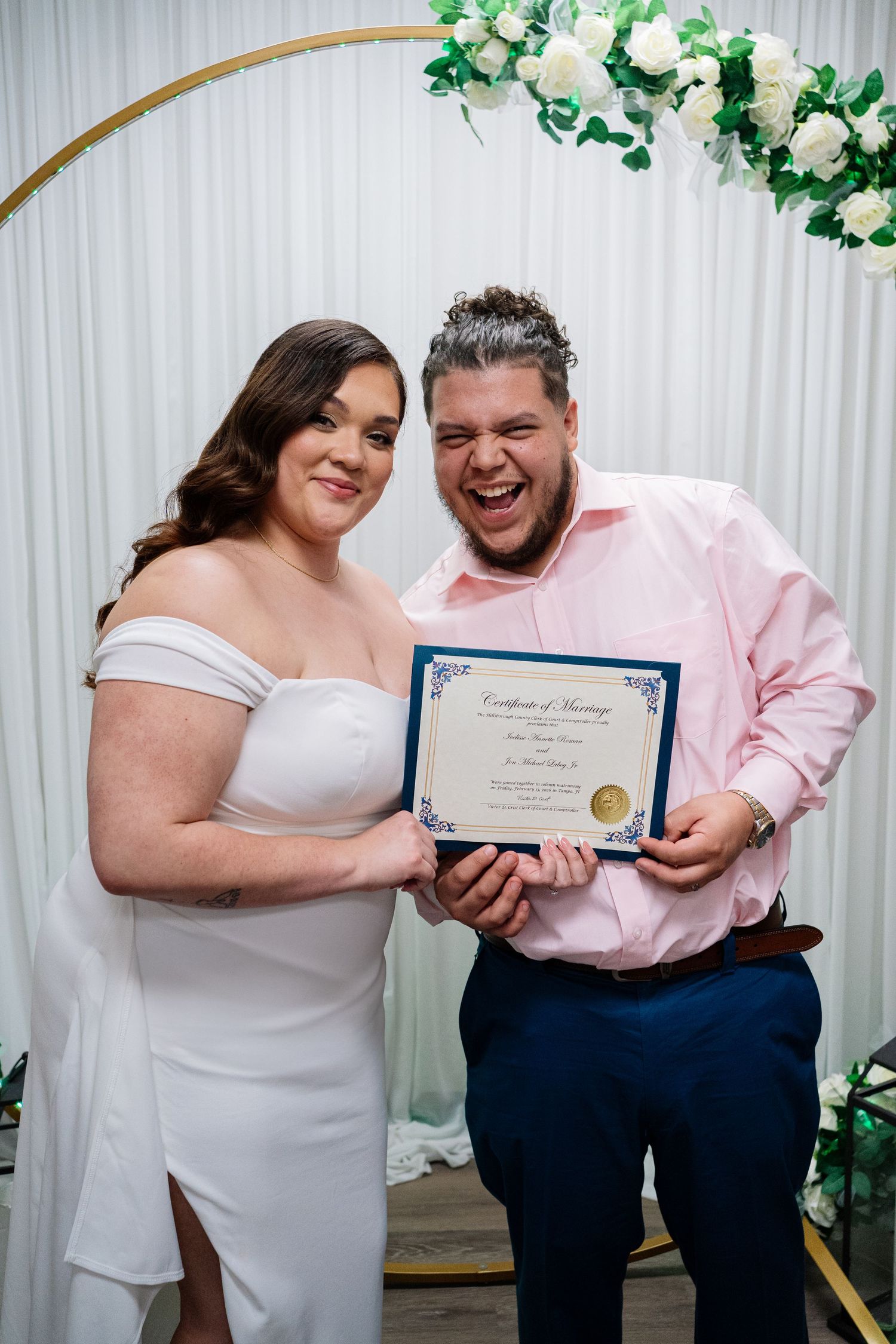 Newlywed couple smiling and holding their marriage certificate in front of a floral arch at their wedding ceremony.