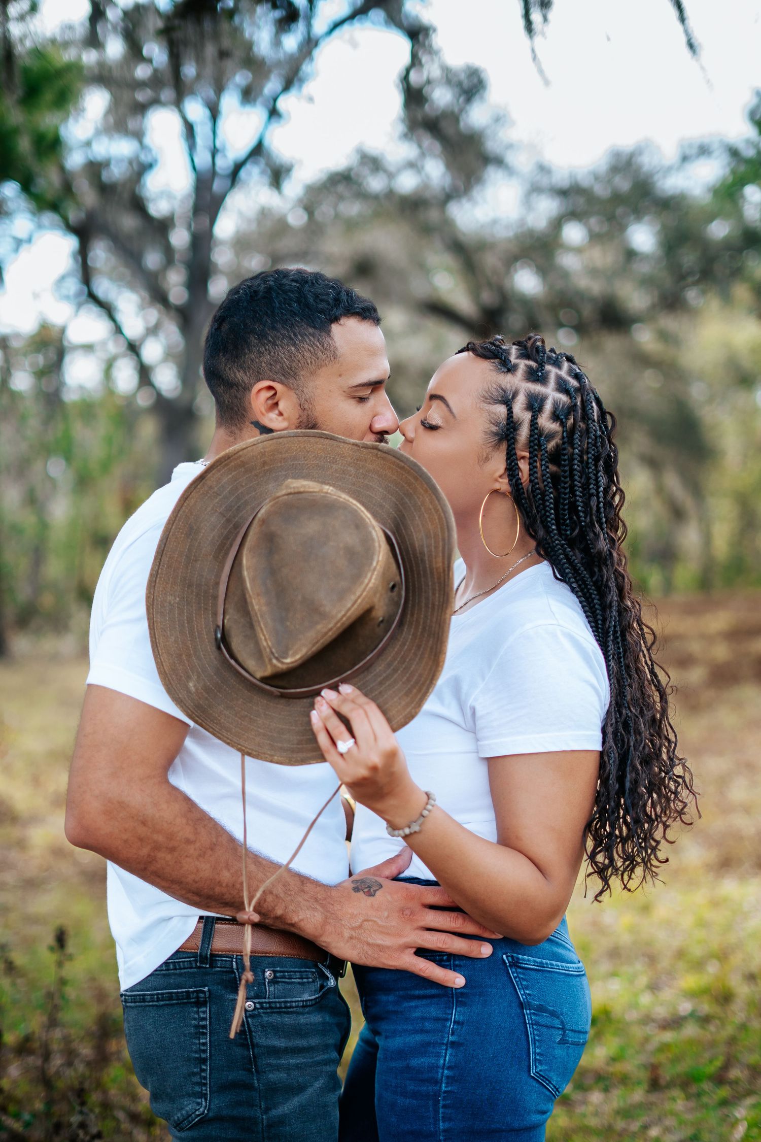 Couple sharing a romantic kiss hidden behind a brown wide-brim hat outdoors in a wooded setting.