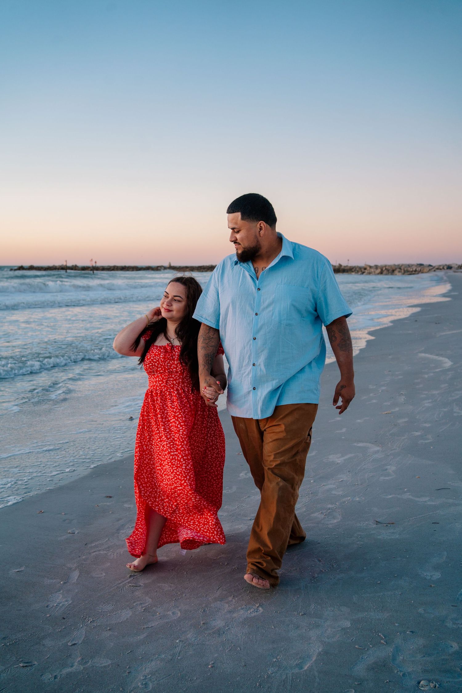 Couple holding hands walking barefoot on beach at sunset, woman in red floral dress, man in blue shirt and tan pants.