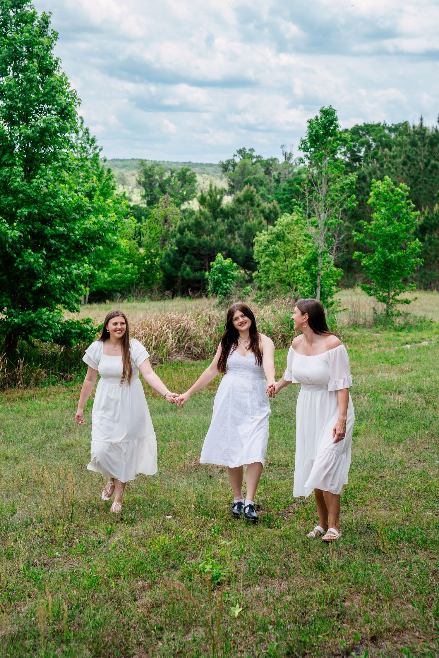 Three women in white dresses hold hands walking through a lush green field with trees and cloudy sky.