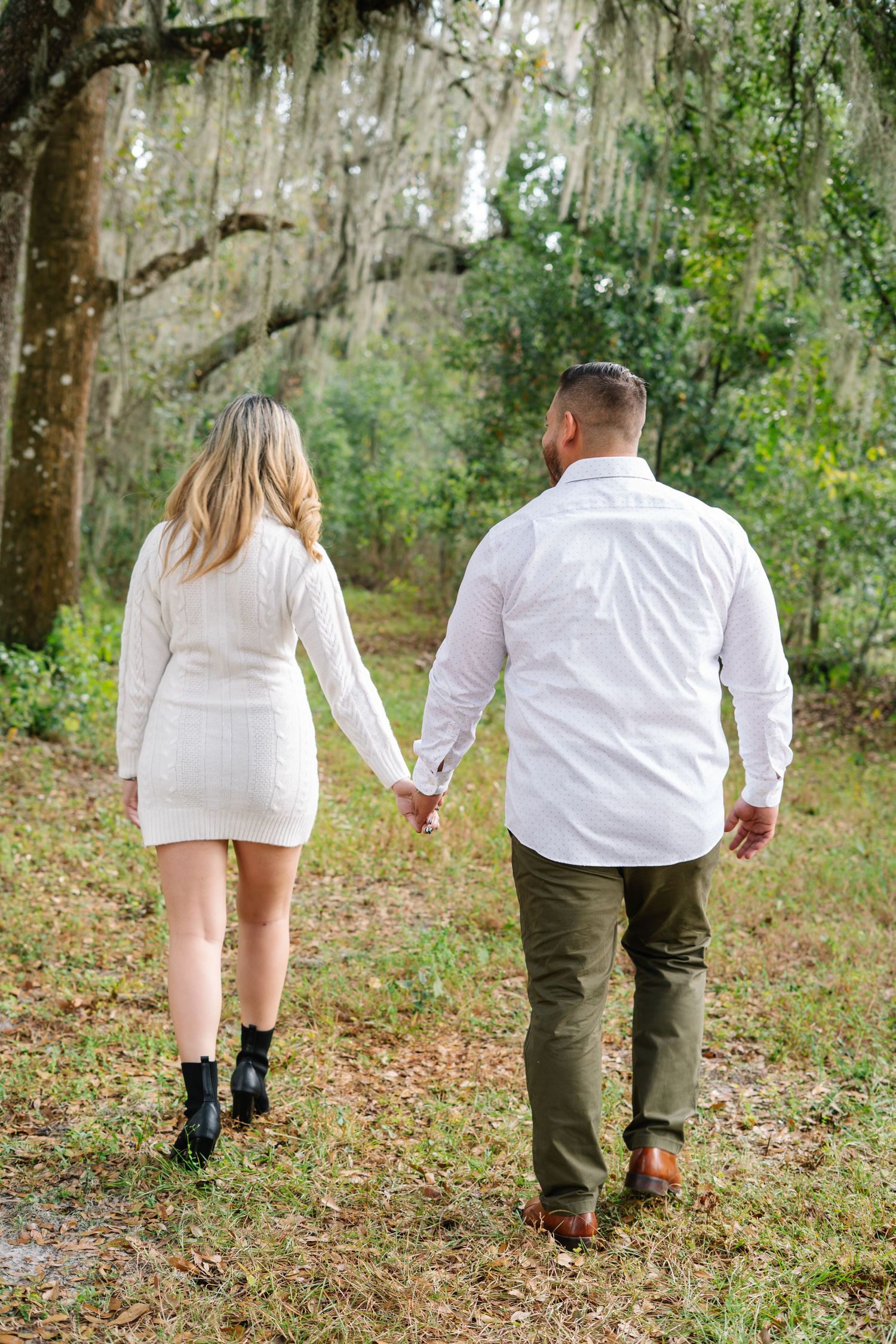 Couple walking hand in hand through a forest wearing coordinated white and olive outfits.