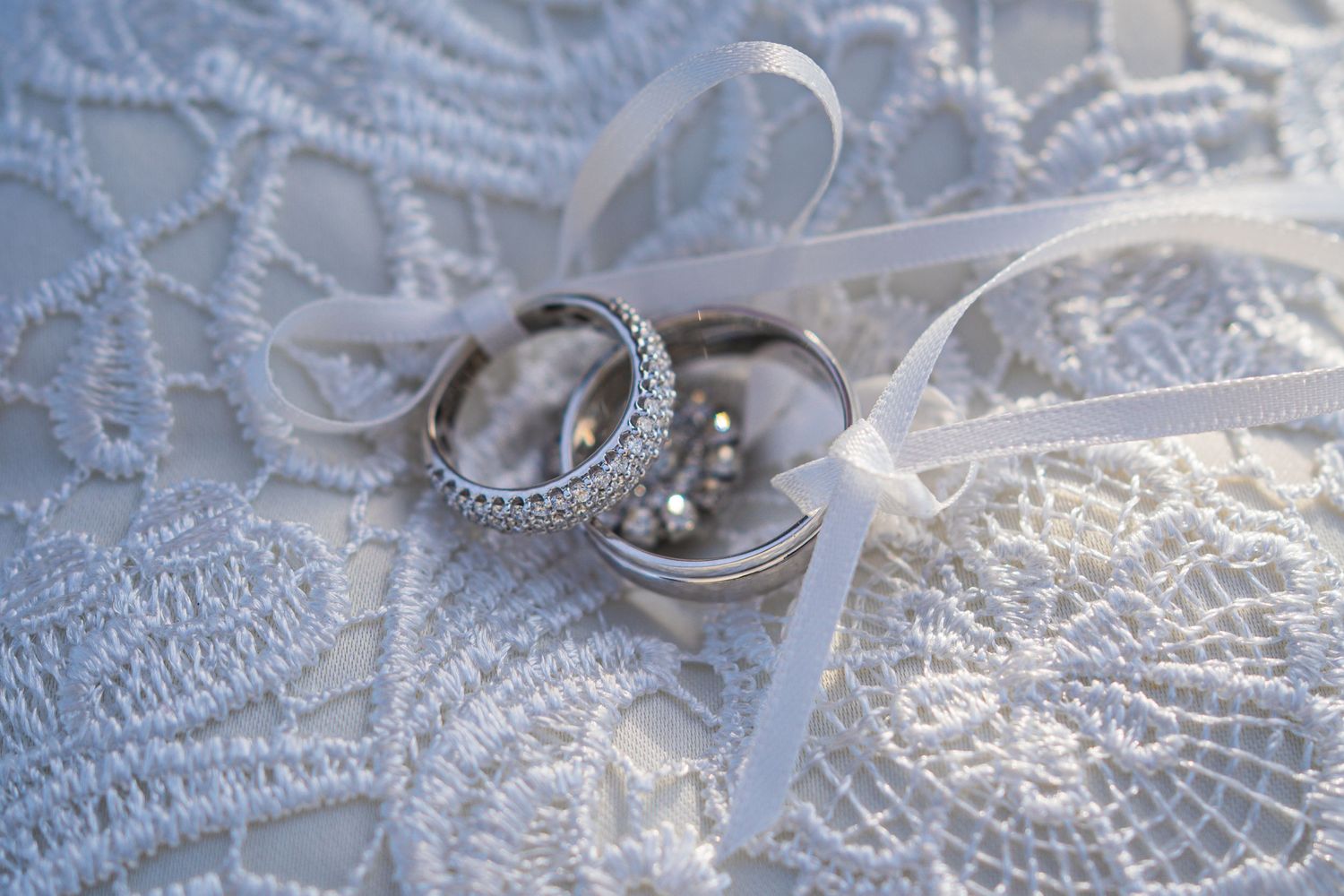 Wedding rings with diamond accents rest on delicate white lace with a sheer ribbon bow.