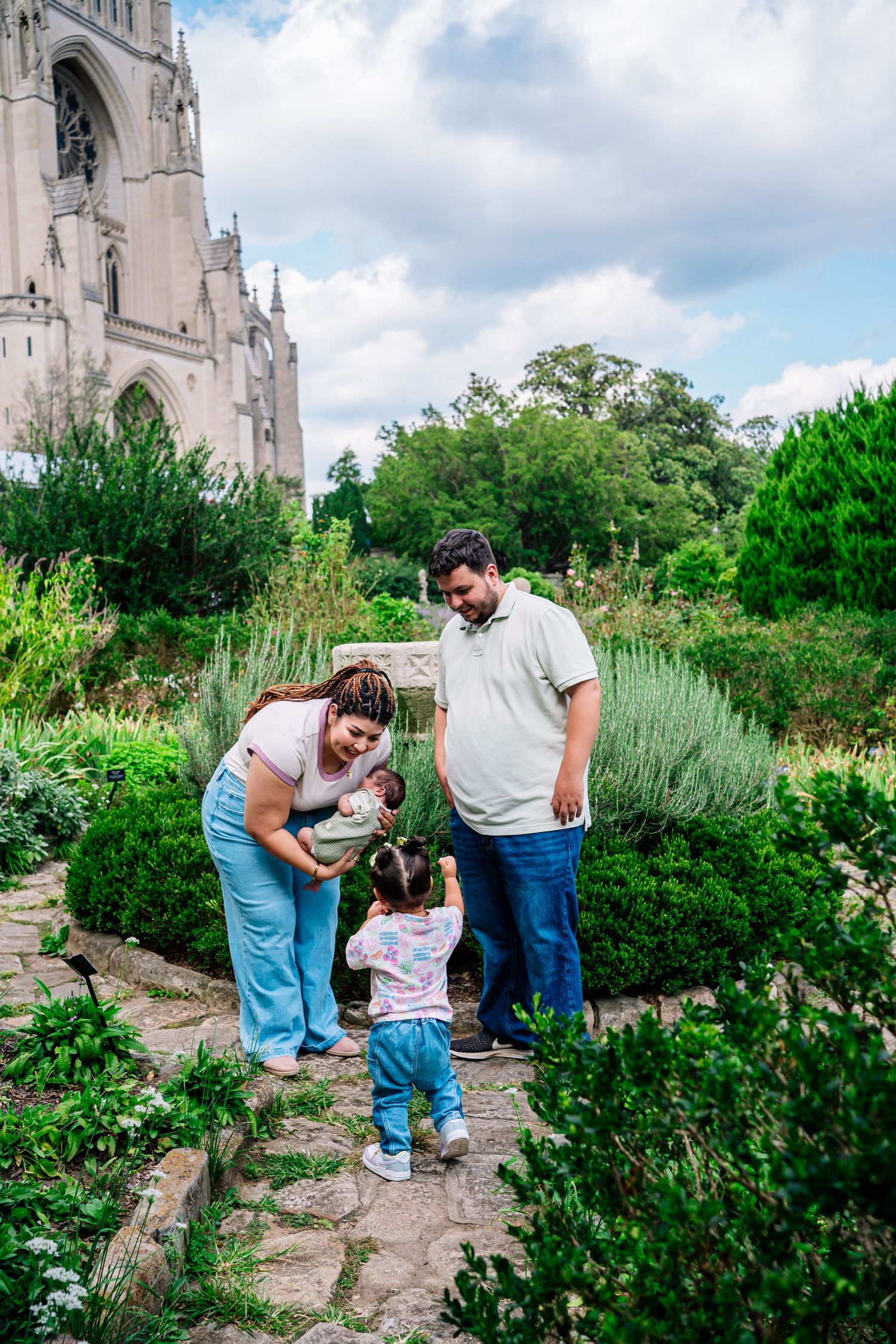 Family with toddler and baby enjoying a garden near a Gothic cathedral on a sunny day.
