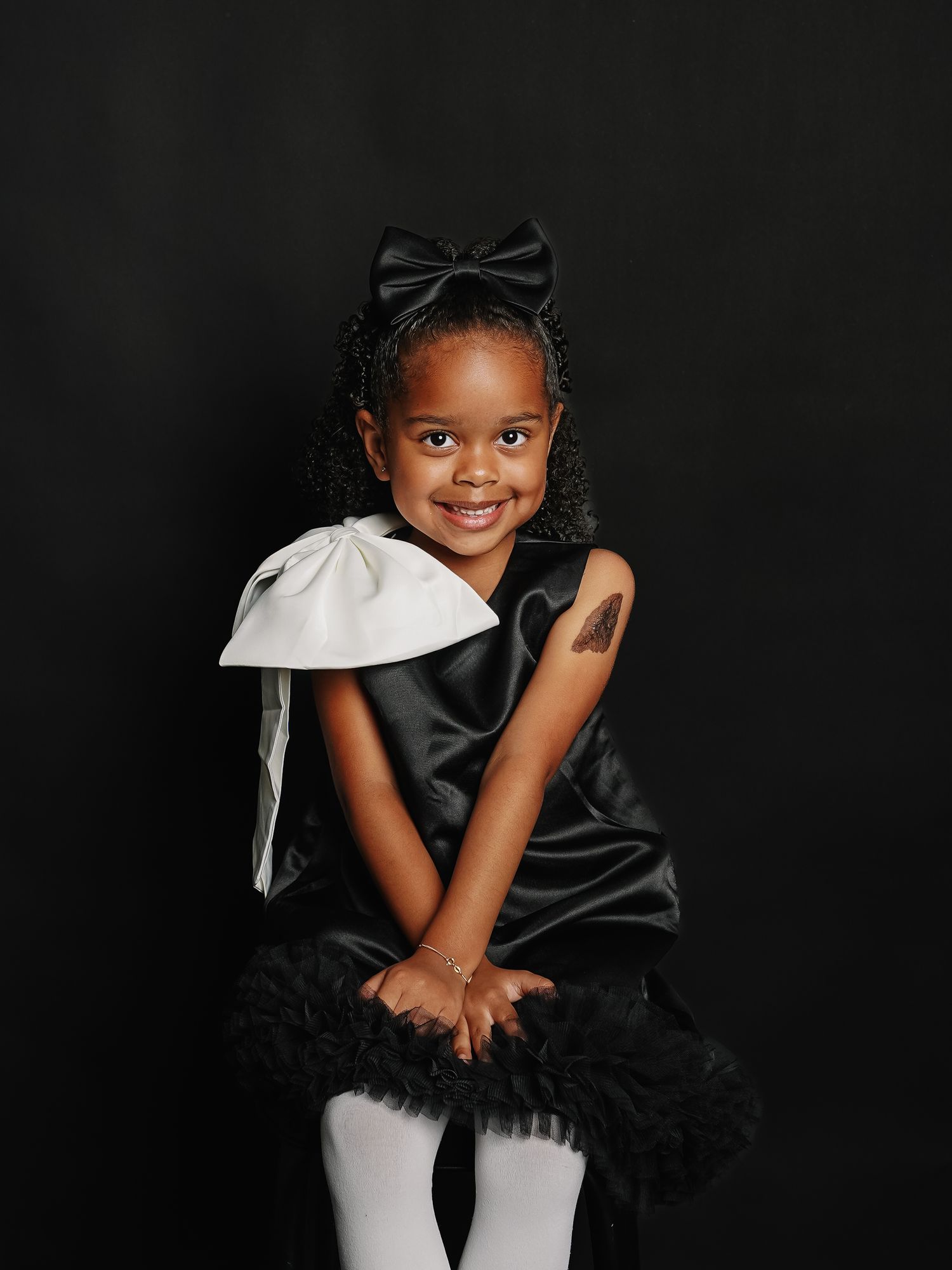 Young Black girl in black satin dress with white bow, black tutu, and hair bow, smiling against dark background.