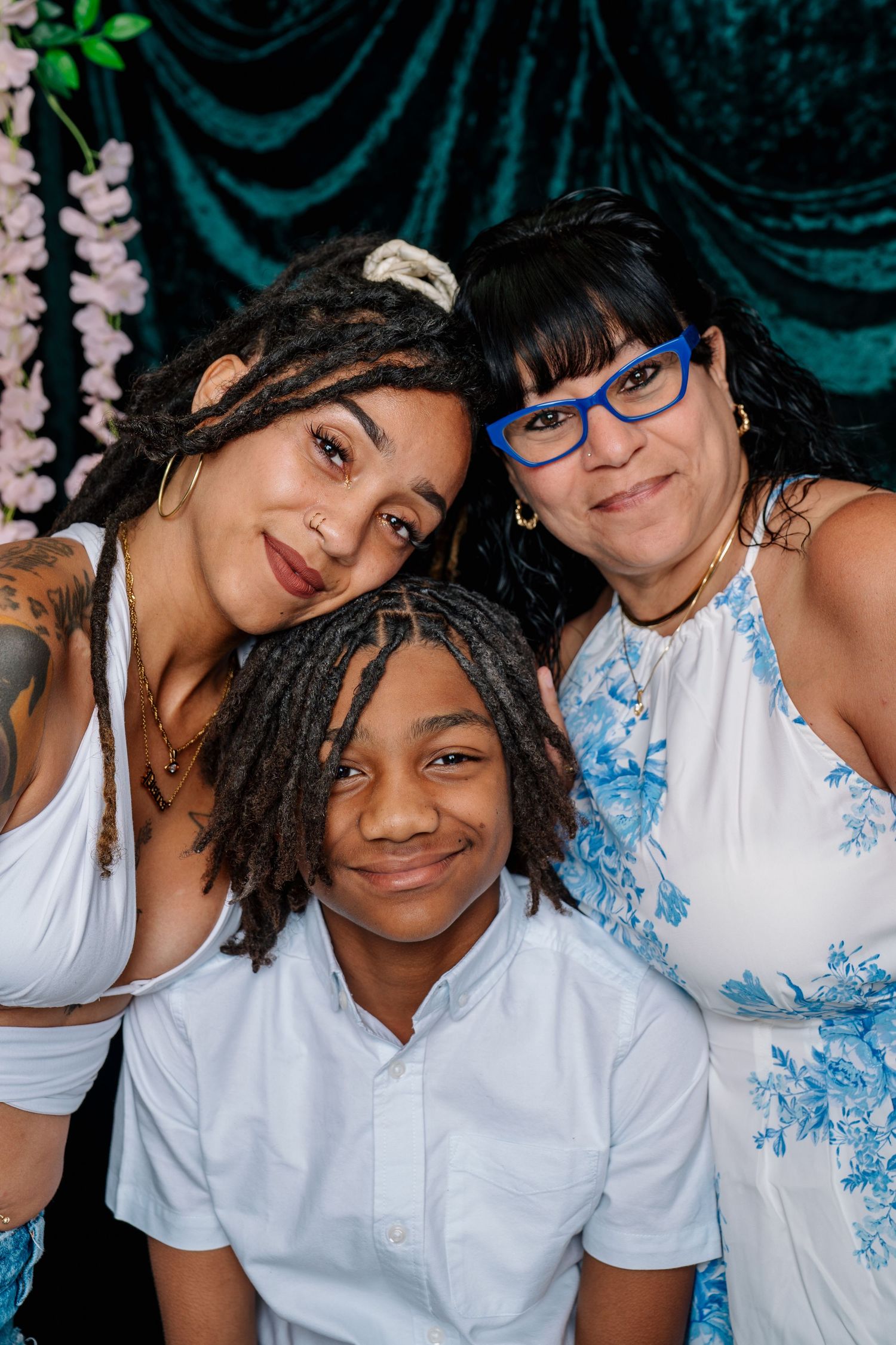 Three people pose together smiling, two women and a young boy, dressed in white against a teal floral backdrop.