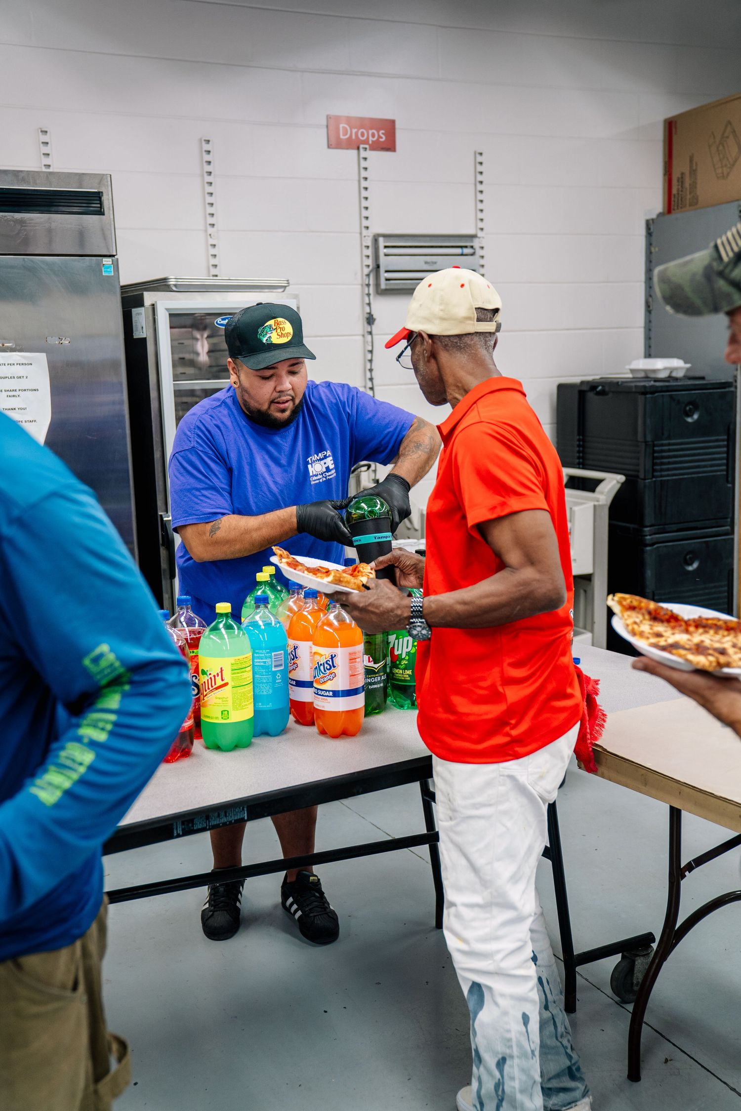 Man in blue shirt serves pizza at a community food event with soda bottles on a folding table indoors.