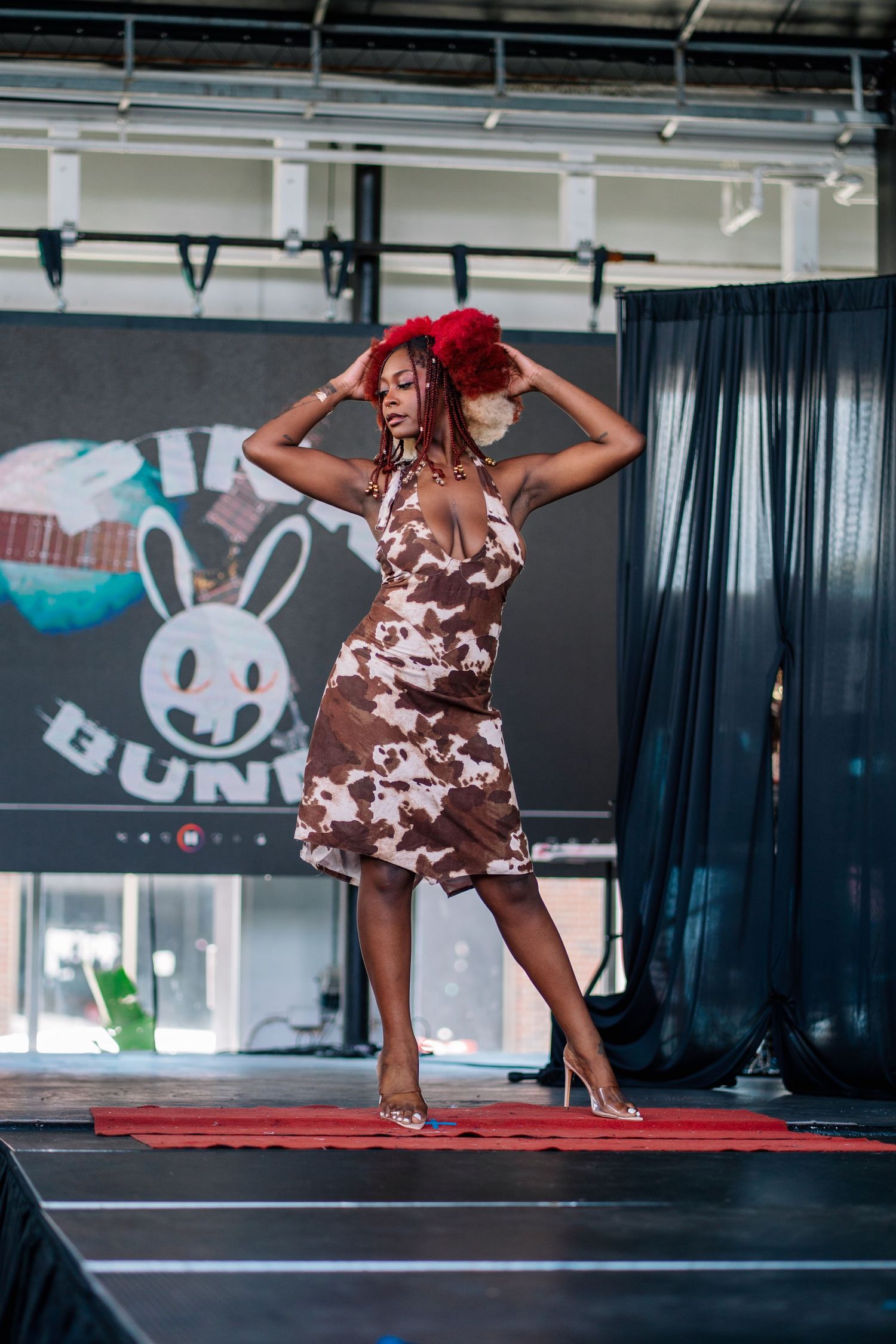Model in cow-print halter dress with red floral headpiece poses on runway stage at fashion show event.