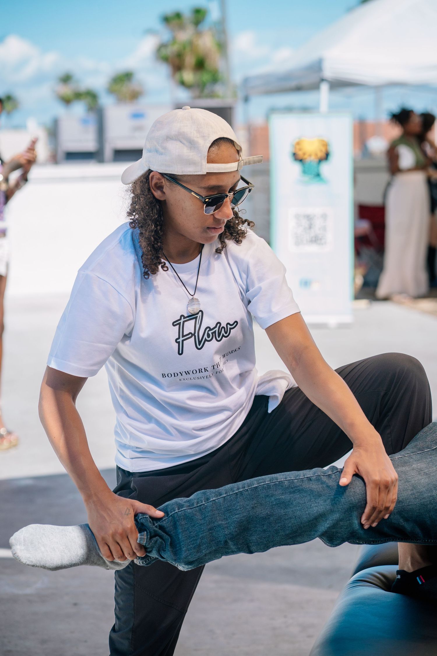 Young man in white Flow t-shirt, snapback cap, and sunglasses dancing outdoors at a sunny event.