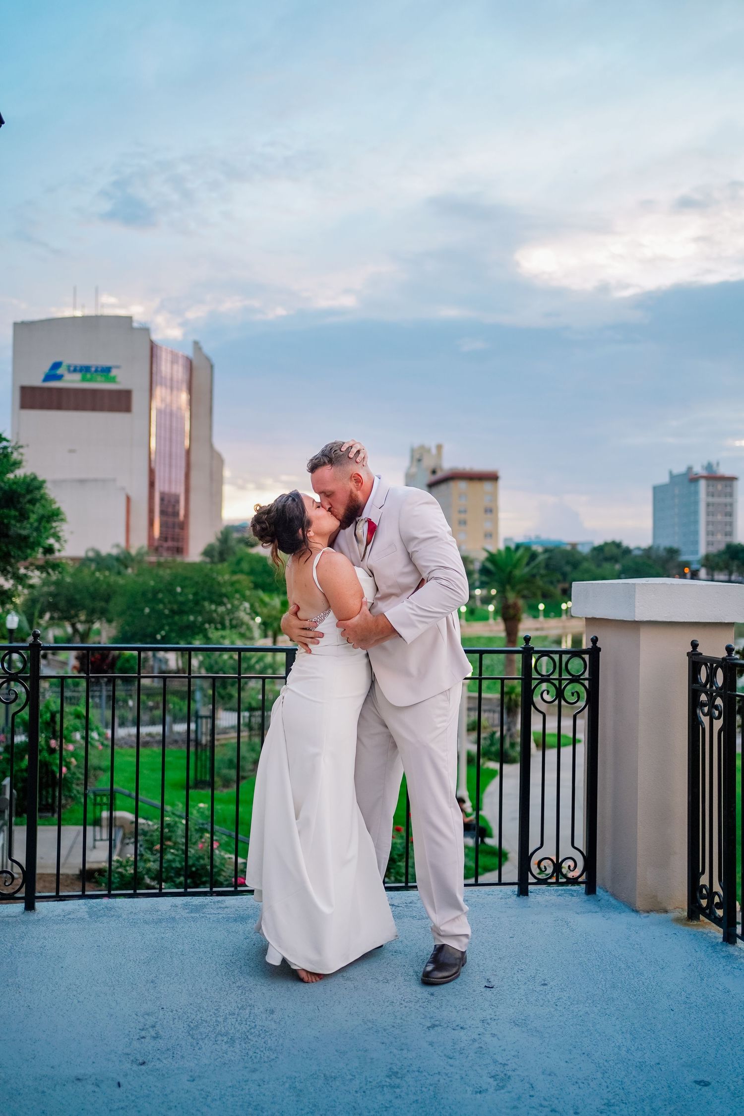 A couple shares a romantic kiss on a rooftop balcony during their wedding, with a city skyline and lush greenery in the background.