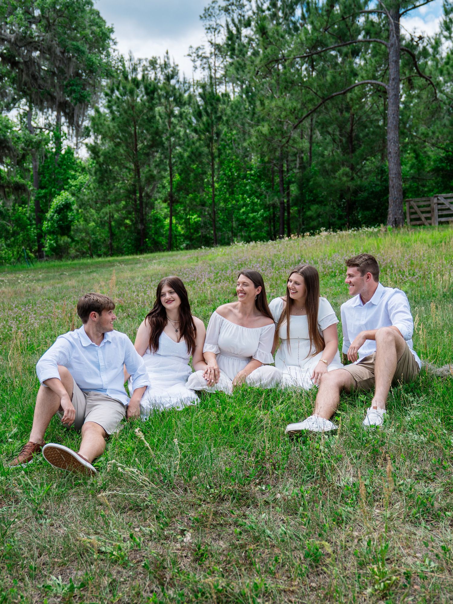 Five young adults sitting and laughing together on a grassy hill, dressed in white and light casual outfits.