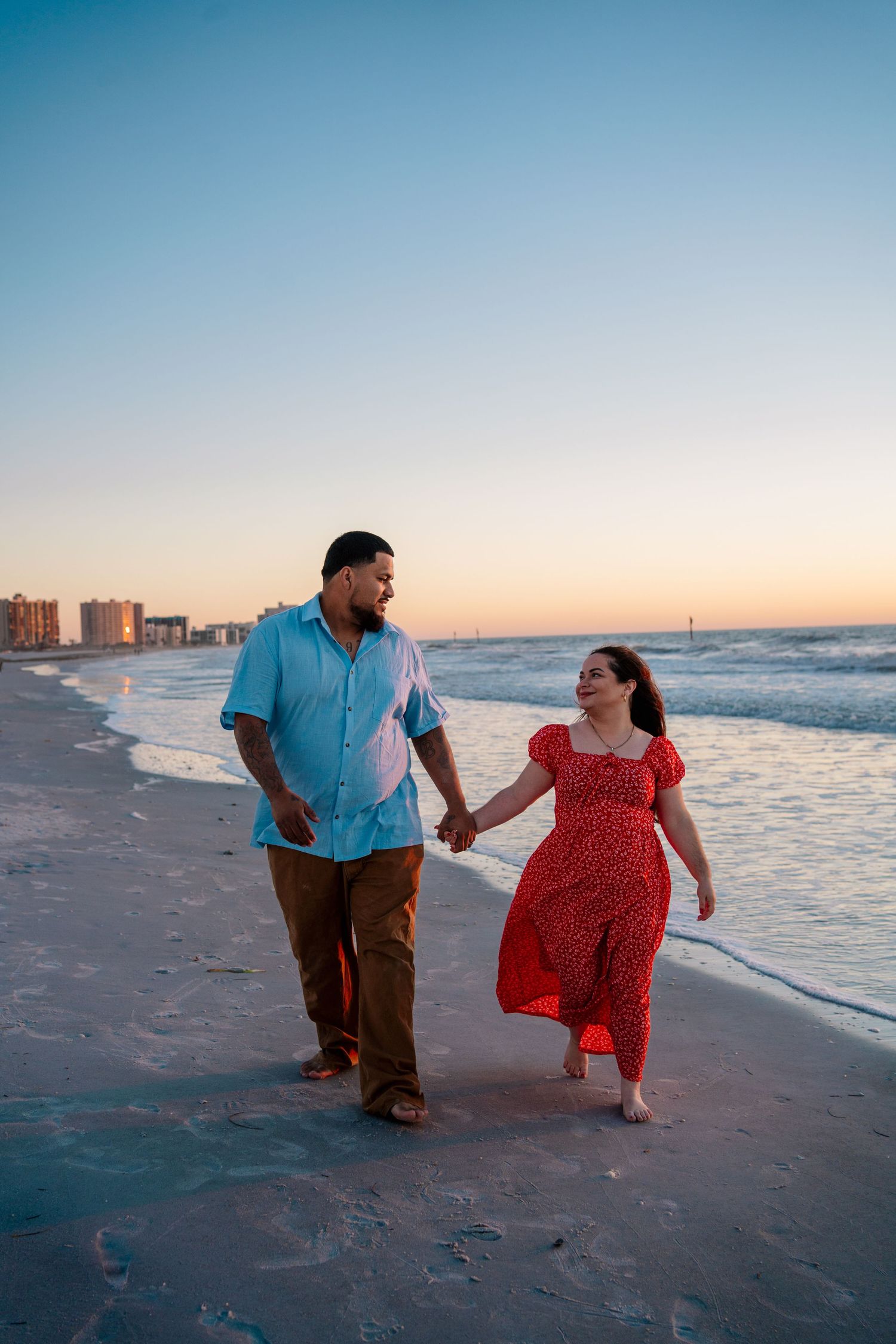 Happy couple holding hands strolling along sandy beach at sunset, woman in red dress and man in light blue shirt.
