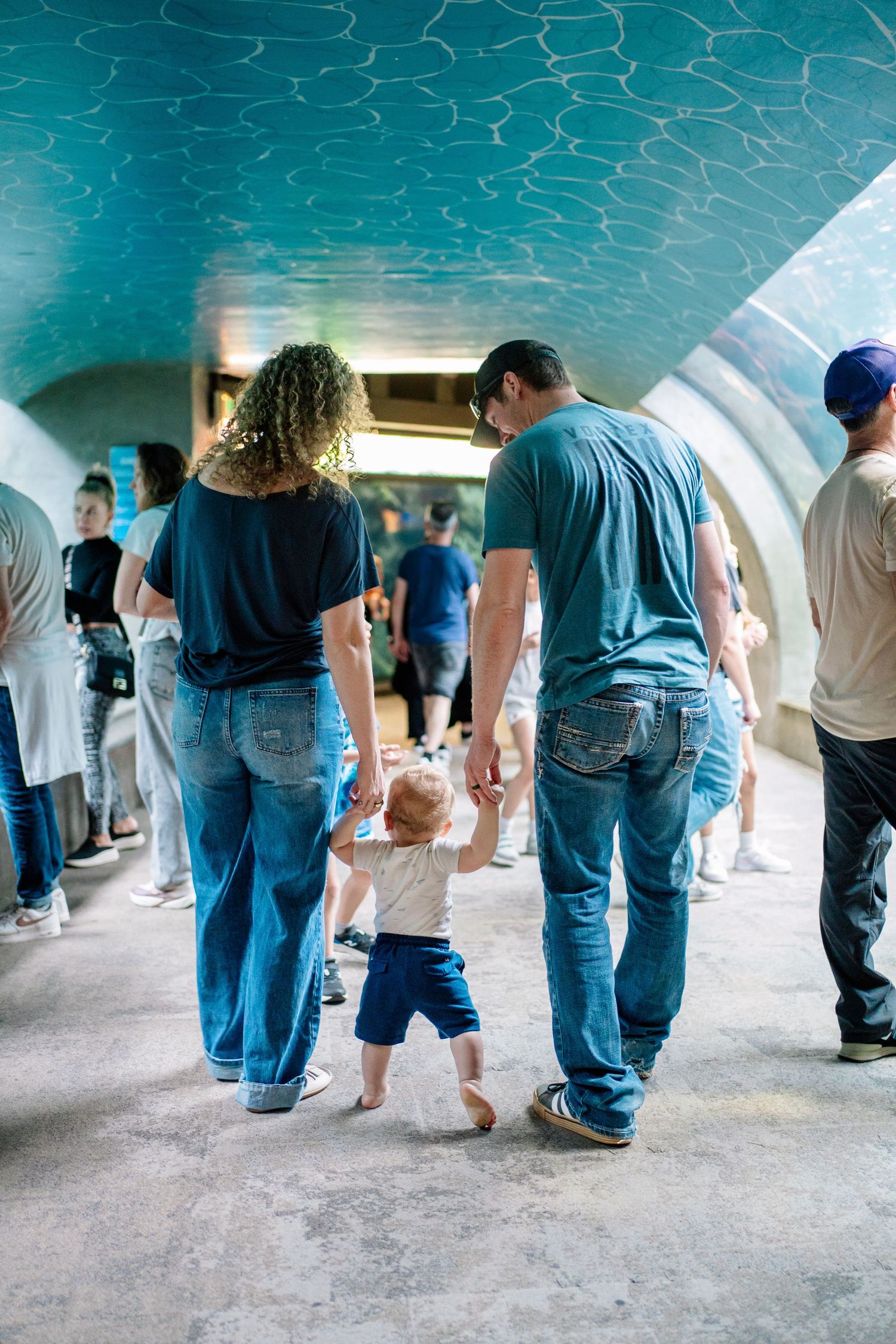 Family with toddler walking through aquarium tunnel, parents holding child's hands, blue curved ceiling overhead.