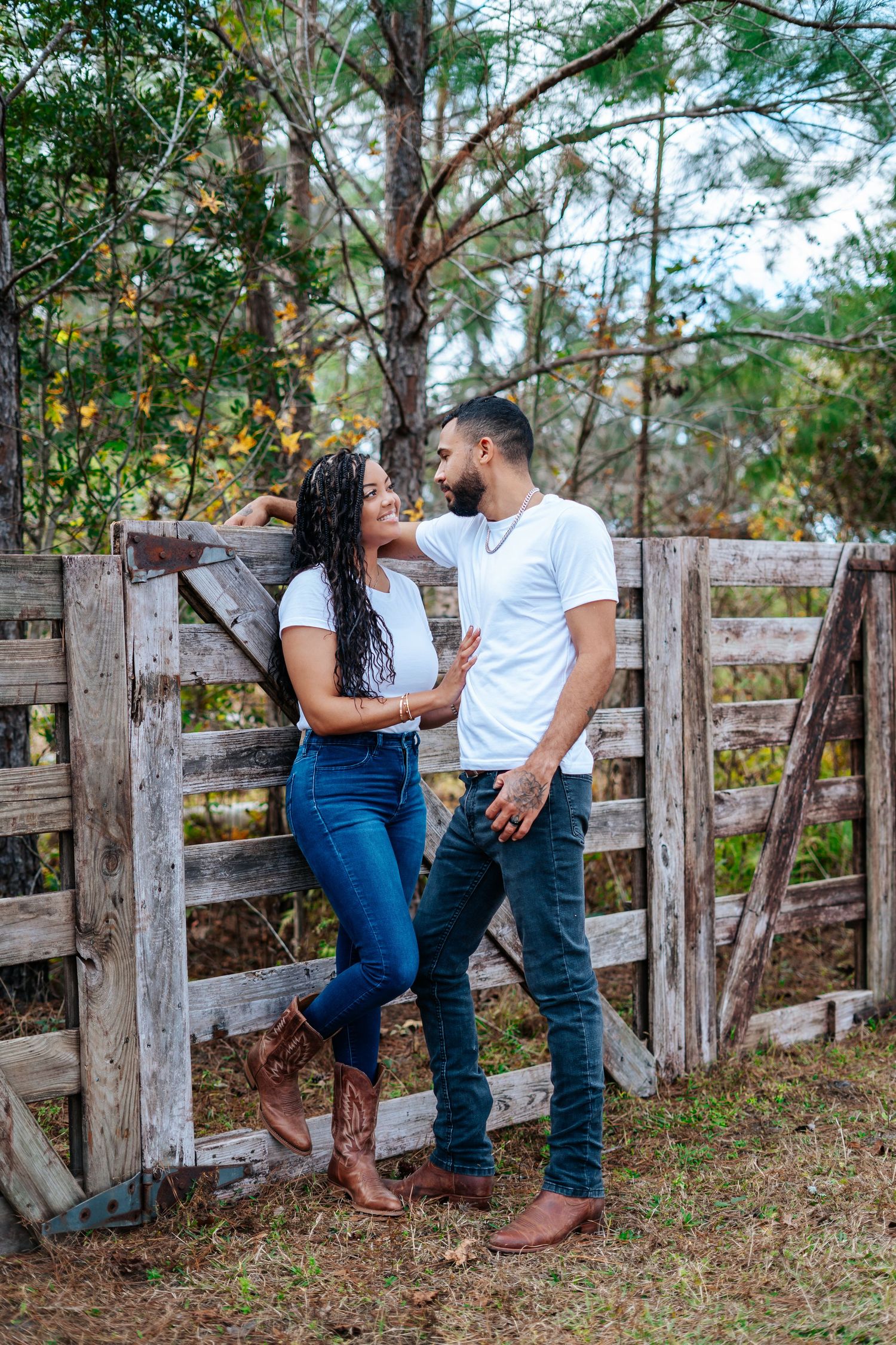 Couple in matching white tees and jeans posing by a rustic wooden fence surrounded by pine trees.