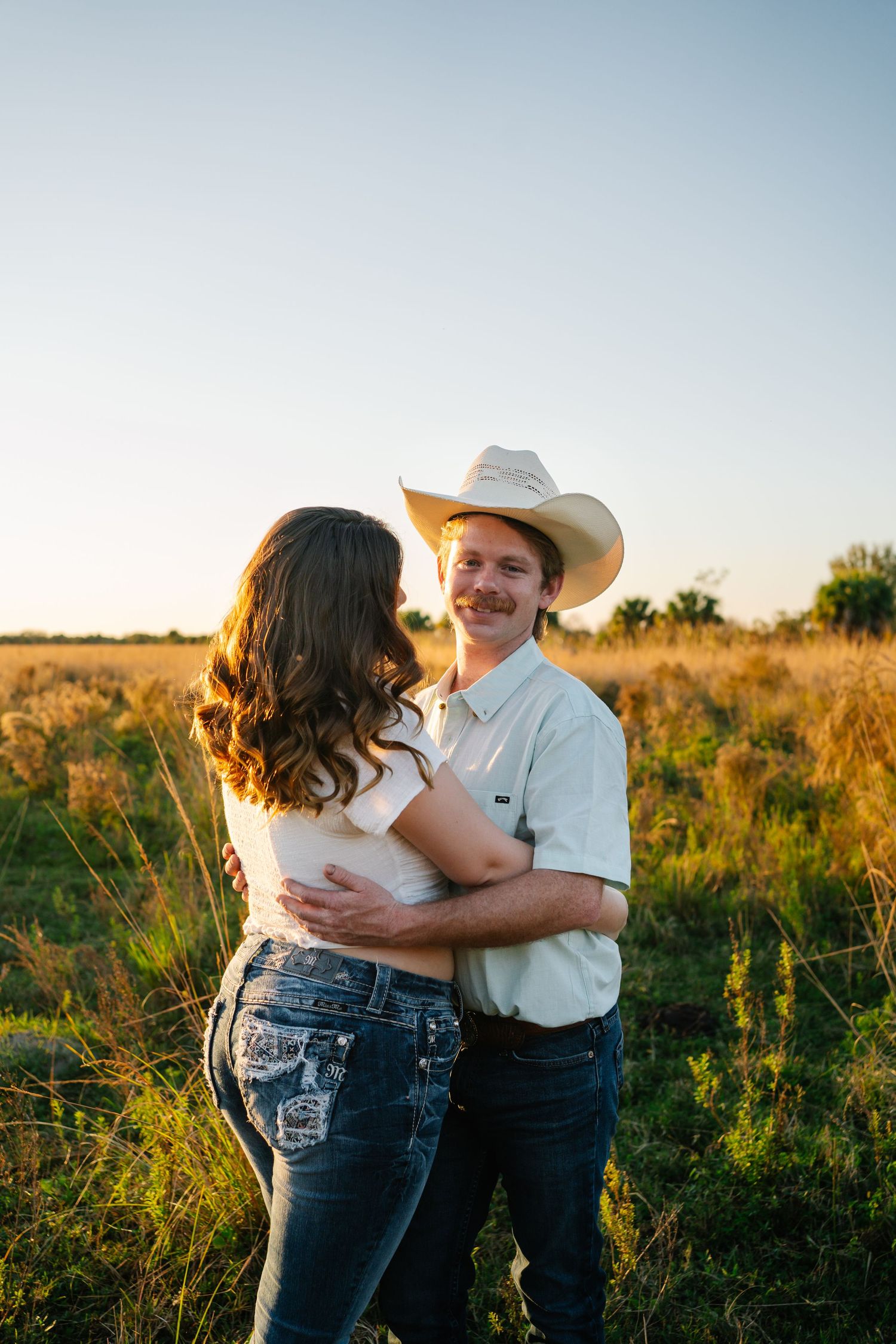 Smiling cowboy with mustache embracing woman in sunny Texas field during golden hour sunset.
