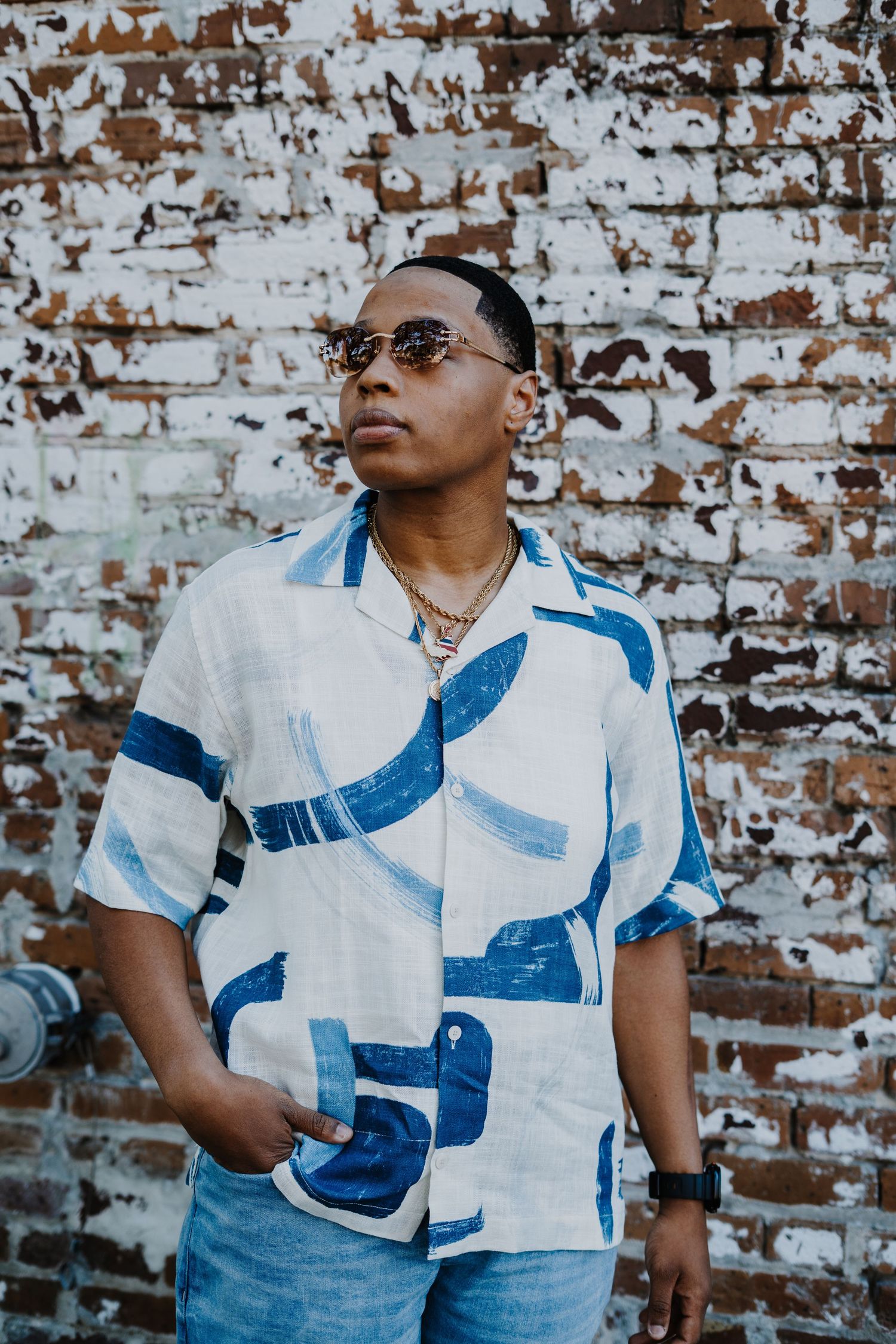 Person wearing blue and white patterned shirt poses against weathered brick wall in artistic portrait series.