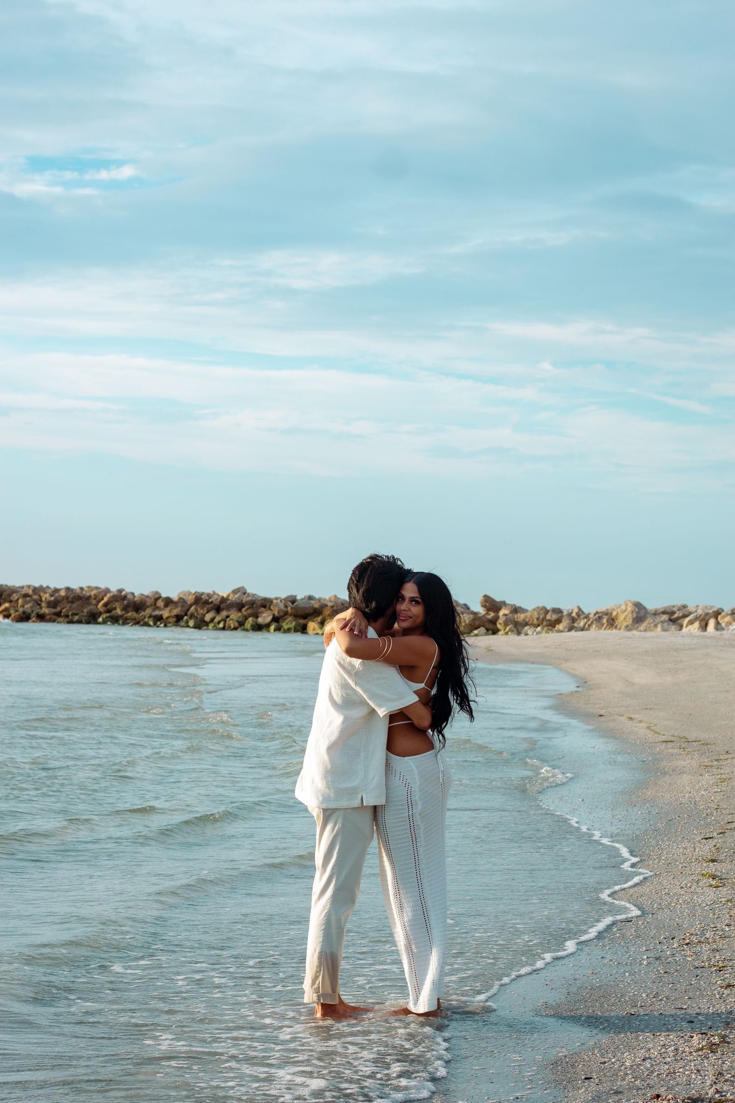 A couple in white outfits shares an intimate moment while walking along a serene beach shoreline.