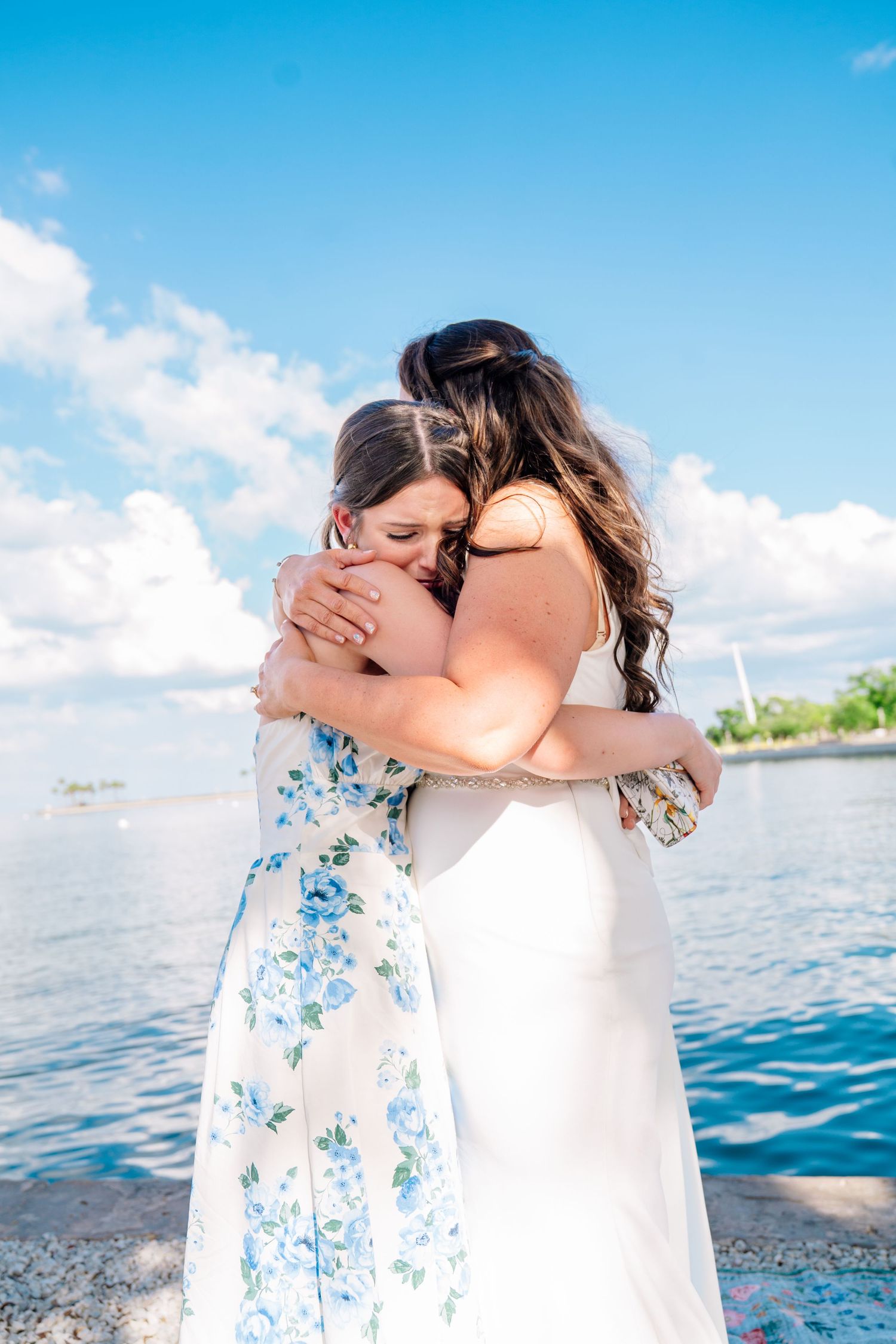 Two women share an emotional hug outdoors near a waterfront on a sunny day, one wearing a white floral dress.